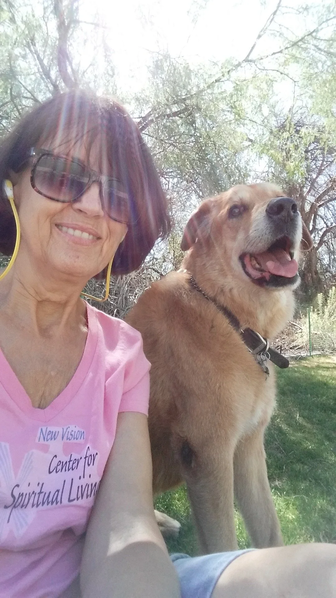 A woman wearing sunglasses and a pink t-shirt taking a selfie outdoors with her dog, a large golden retriever, on a sunny day with trees in the background.