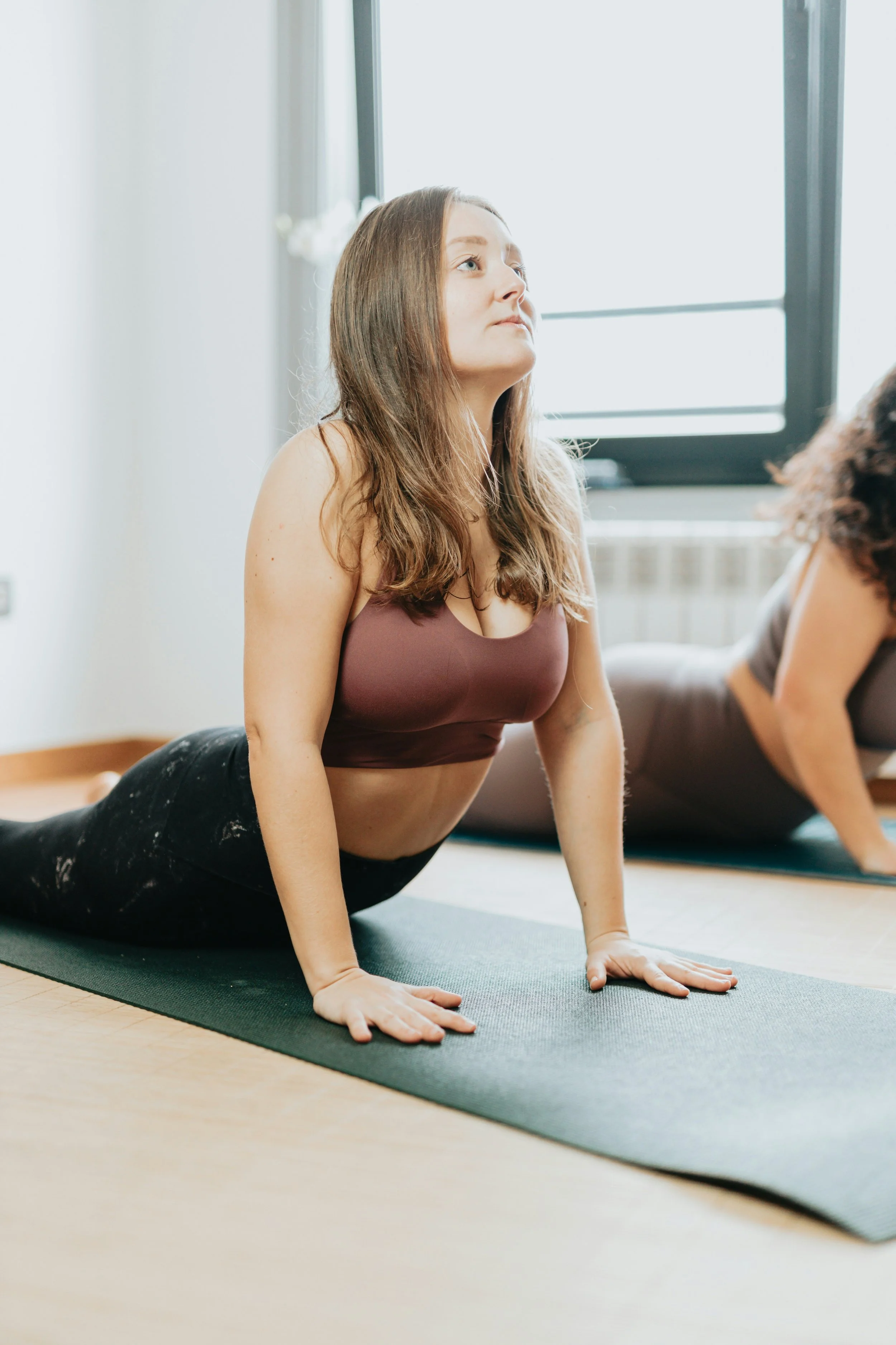 Une jeune femme fait du yoga, position en inspirant, sur un tapis dans une pièce lumineuse avec fenêtre