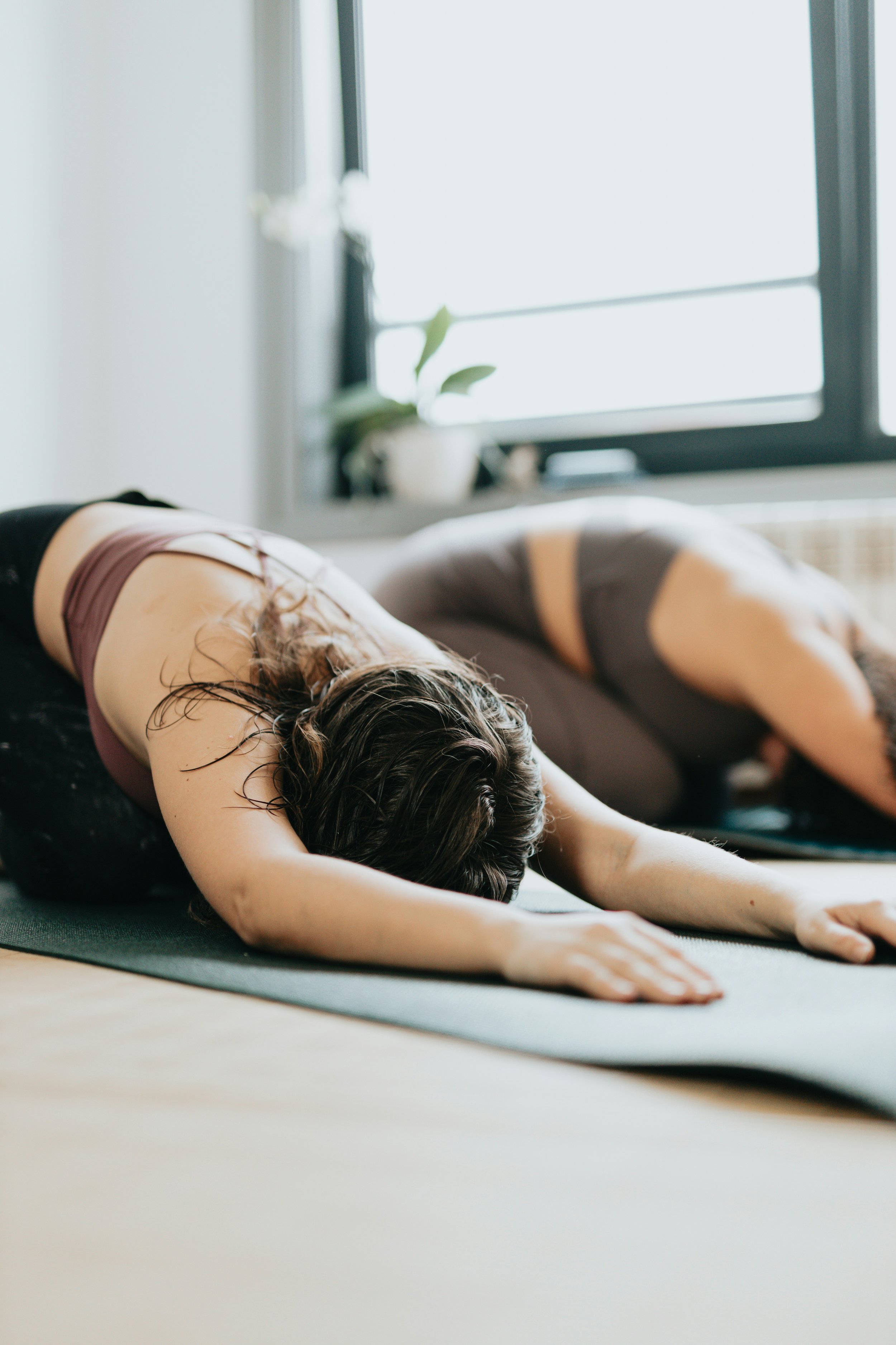 Une personne faisant une posture de yoga sur un tapis dans une pièce lumineuse avec une fenêtre en arrière-plan et une plante en pot sur le rebord de la fenêtre.