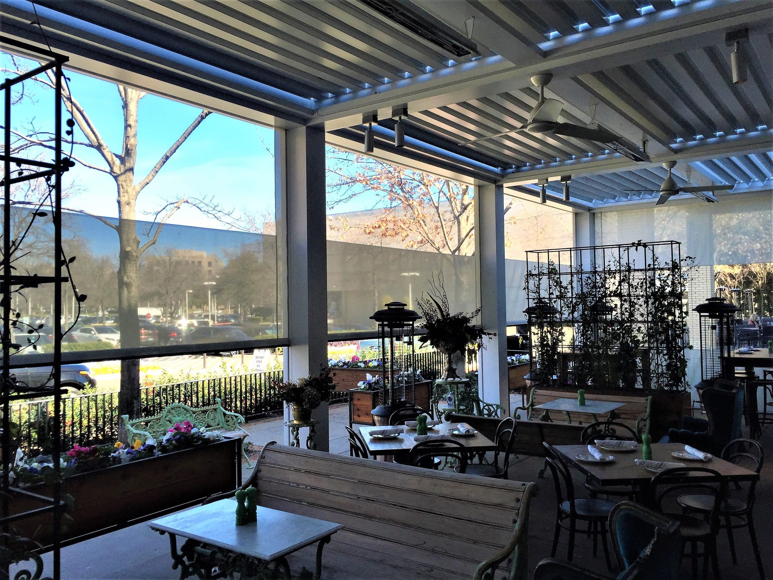 Indoor restaurant patio seating area with tables, chairs, plants, and large glass windows showing trees and parking lot outside.
