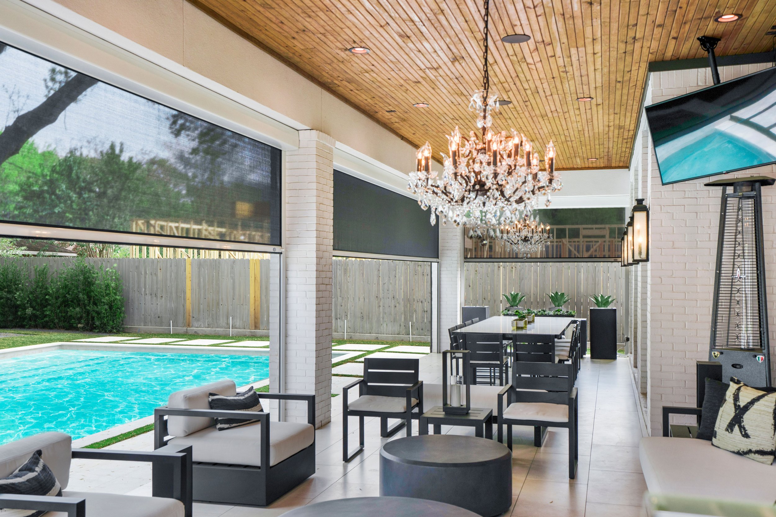 Indoor patio protected by retractable screens with modern furniture, chandeliers, a pool outside, and a flat-screen TV mounted on the wall.