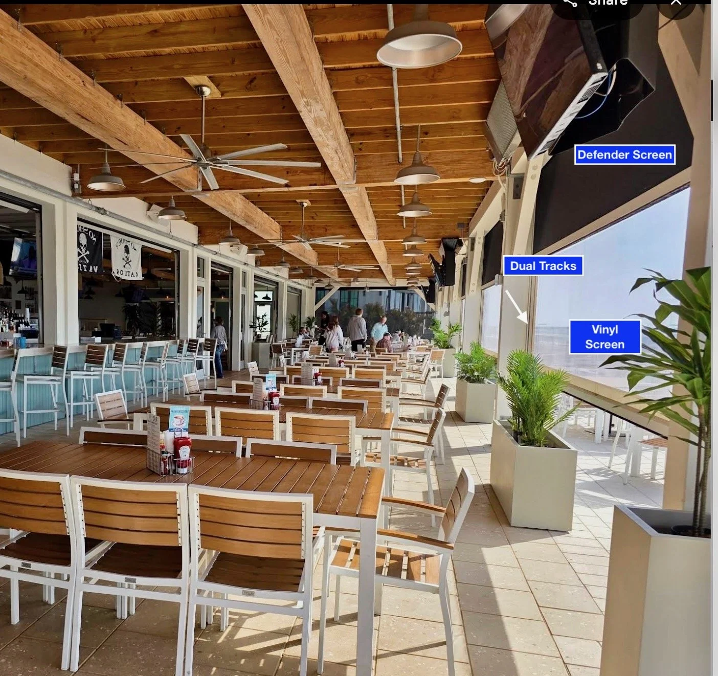 Indoor dining area with wooden ceiling beams, ceiling fans, and pendant lights. There are several tables and chairs arranged for customers. Blue signs label various electronic screens near the windows, including a defender screen, dual tracks, and a vinyl screen. Potted plants are placed along the window area, and there are a few people standing and walking.