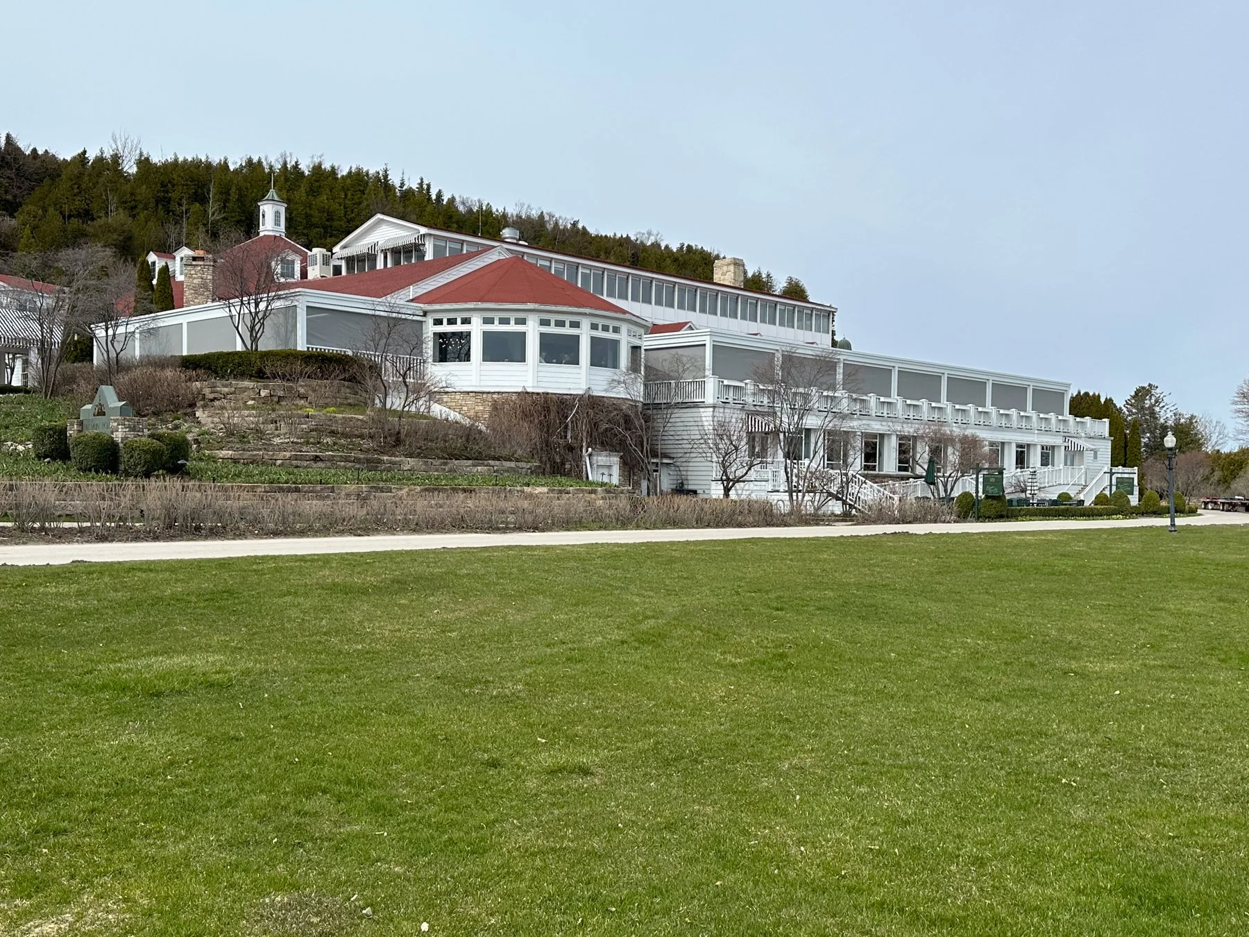 A large white house with a red roof, multiple levels, and extensive porches, situated on a hillside with a grassy lawn in the foreground and a wooded hill in the background.