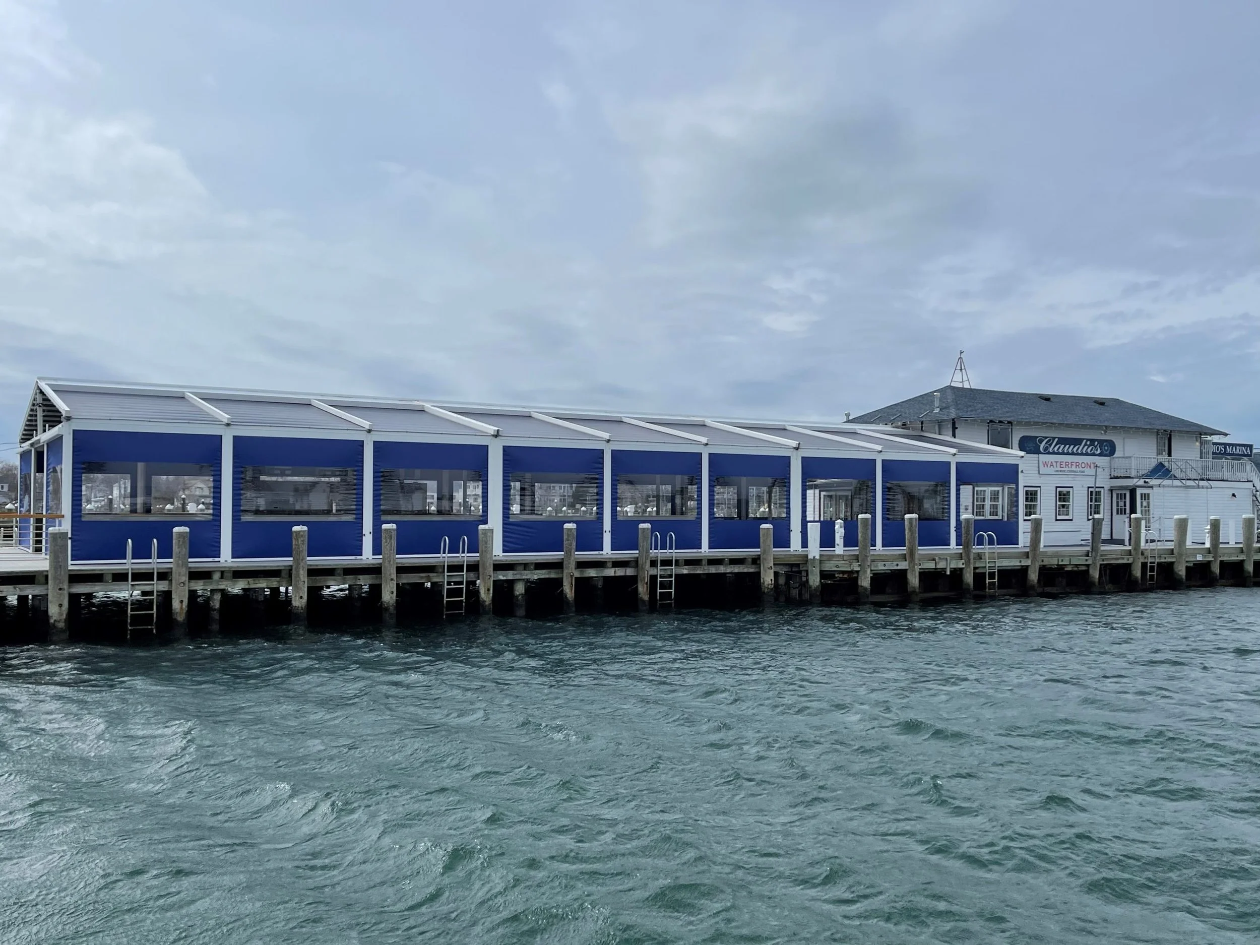 A waterfront restaurant with a blue and white exterior, situated along a dock on the water. The sky is cloudy with some patches of blue visible.