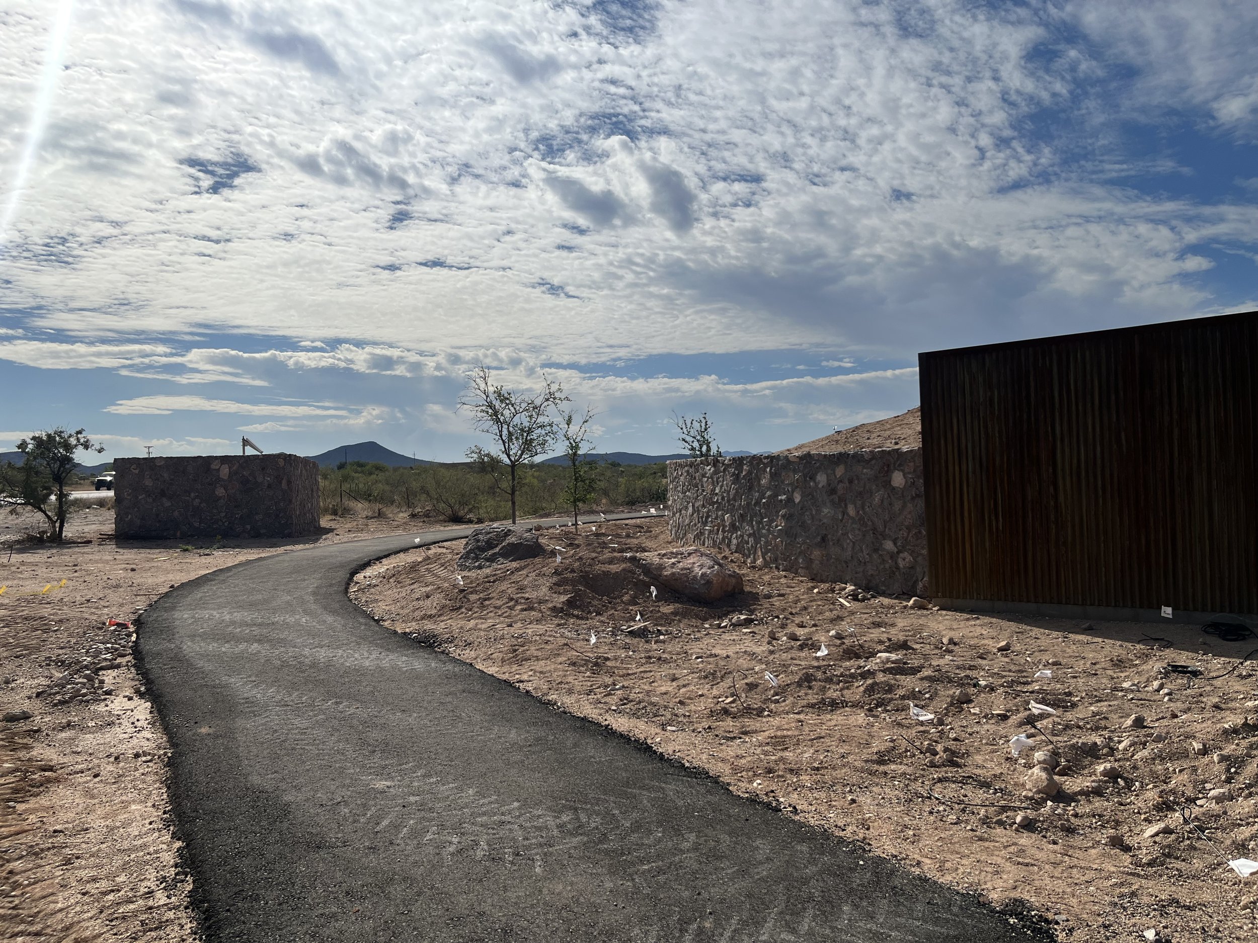 A winding paved path in a desert landscape with small trees, rocks, and construction barriers, under a partly cloudy sky with mountains in the distance.
