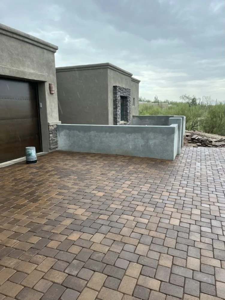 A residential driveway with interlocking bricks, a solid concrete wall, and a modern building with a garage door and stone accents. Overcast sky with visible rain.