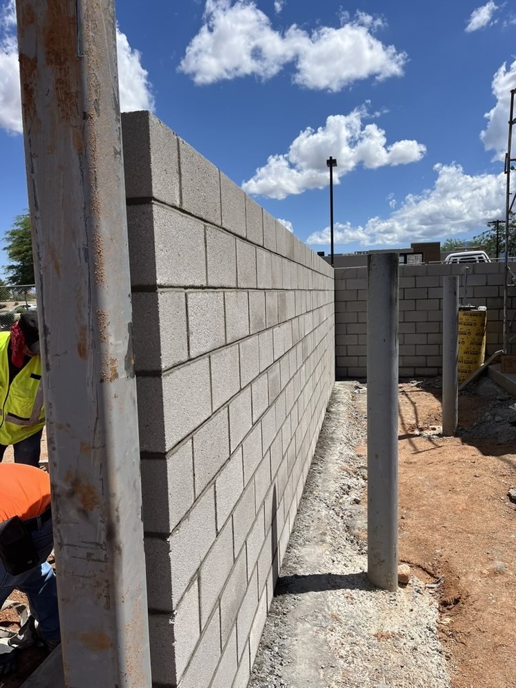Construction site with a partially built cinder block wall, metal posts, scaffolding, and construction workers under a partly cloudy sky.