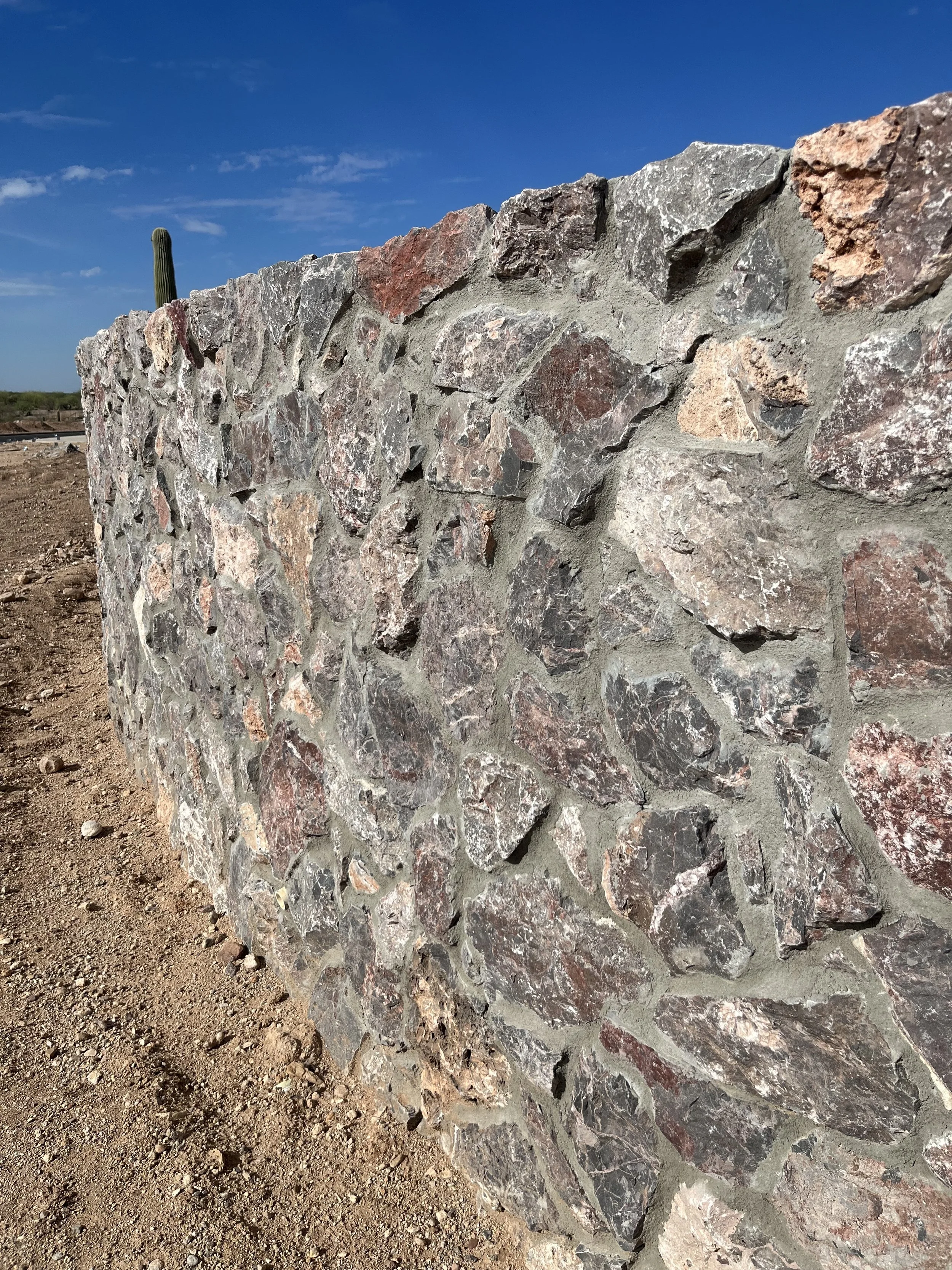 A close-up of a stone wall made of various types of rocks, with a desert in the background and a clear blue sky with a few clouds.