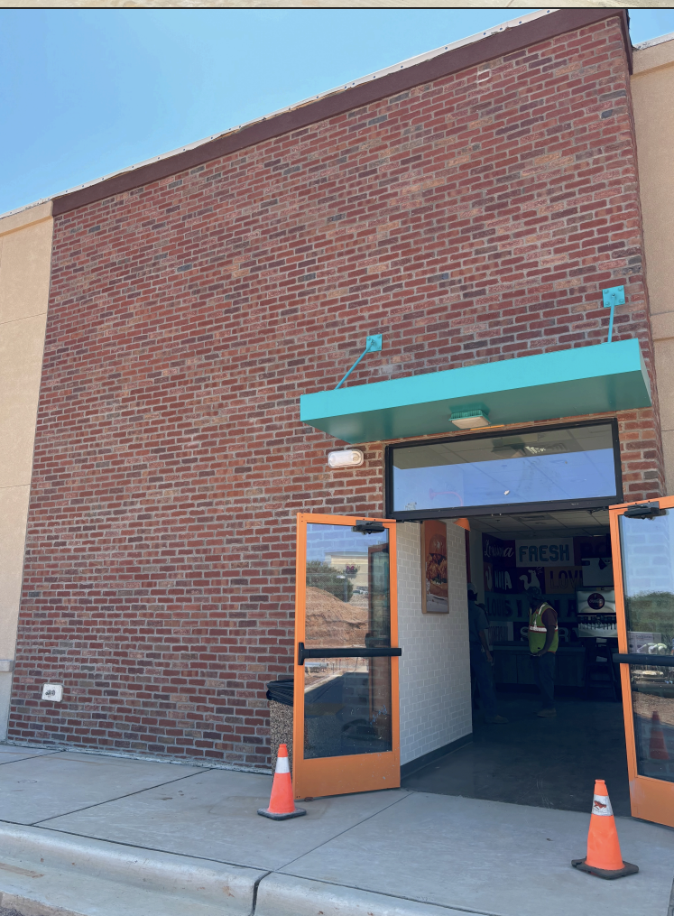 Entrance of a building with orange-framed glass doors, a green awning, and a brick facade, with construction cones outside.