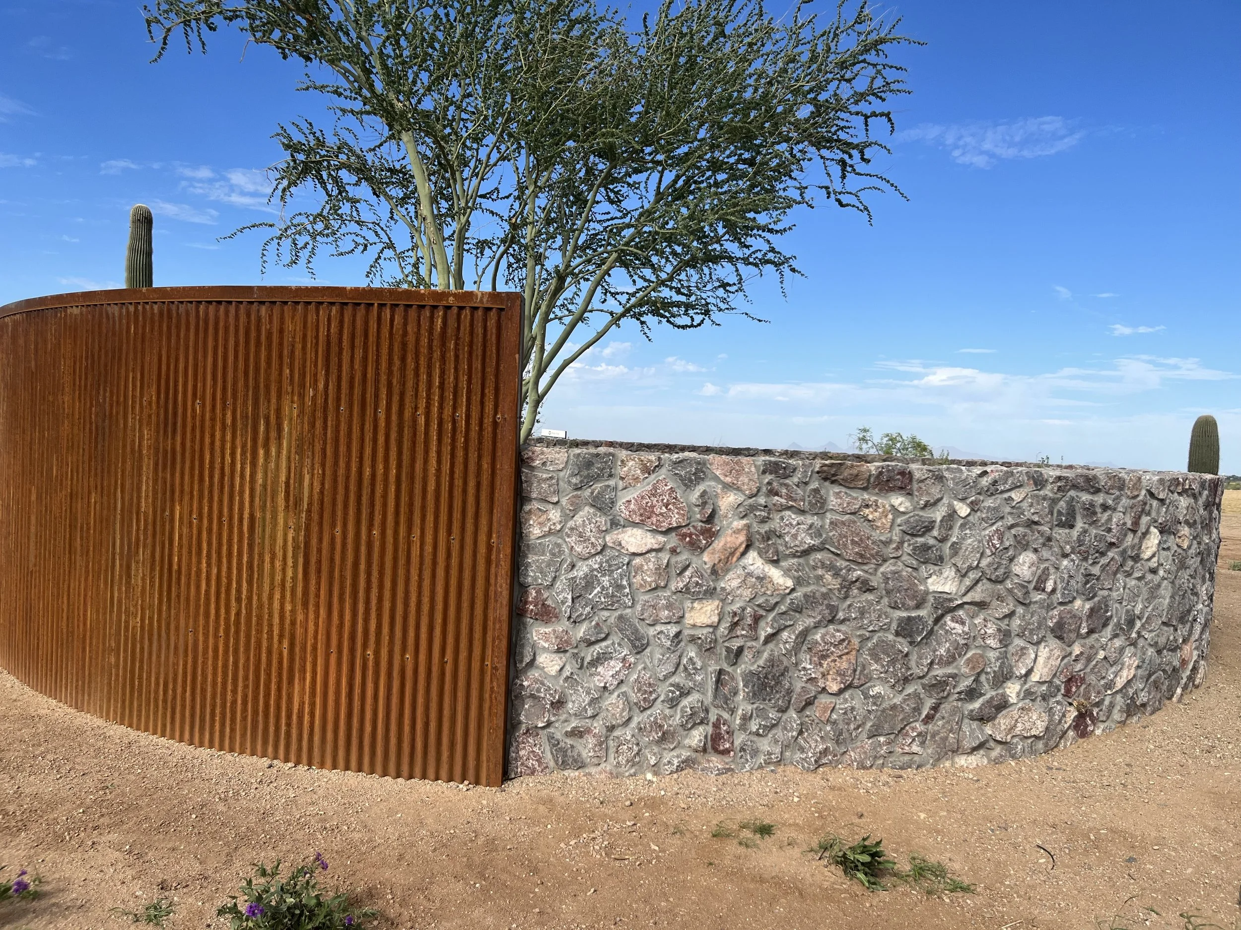 A metal privacy screen with a rust-colored finish, a stone wall, a tall tree with sparse leaves, and a cactus in the background against a blue sky with scattered clouds.