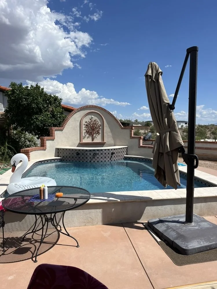 Backyard swimming pool with a fountain featuring a decorative tree design on the wall behind the pool. A white swan-shaped inflatable float is in the pool. There is a round mesh outdoor table with some toys and drinks on it, and a large closed patio 