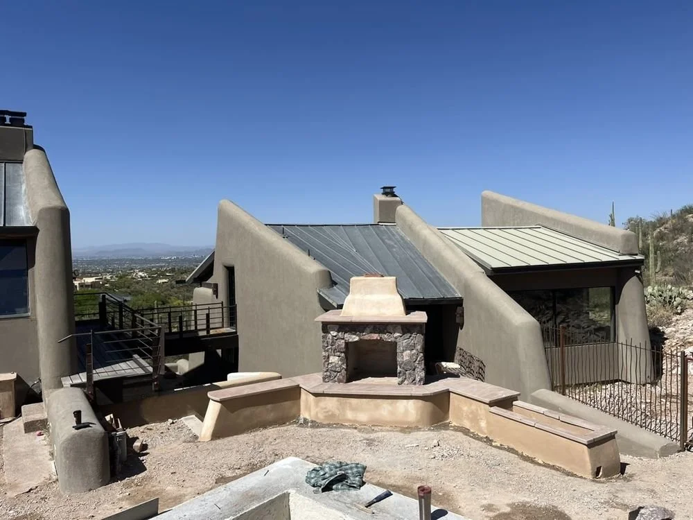 A modern house with a unique, sculptural architectural design featuring angular walls, metal roofing, and an outdoor fireplace, situated in a desert-like landscape under a clear blue sky.