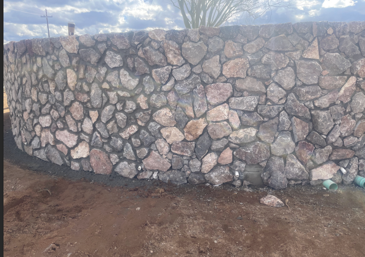 A curved stone wall made of irregularly shaped rocks with visible mortar, set on a ground of dirt, with a clear sky and a tree in the background.