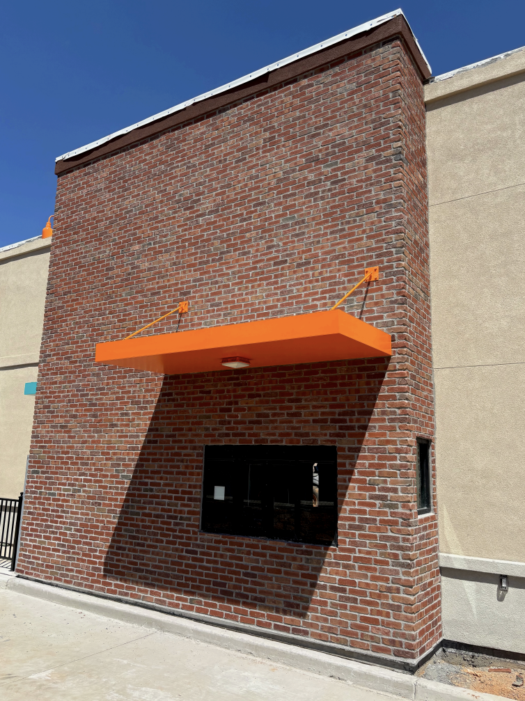 A brick building facade with an orange awning and a small window, with a shadow cast by the awning on the brick wall.