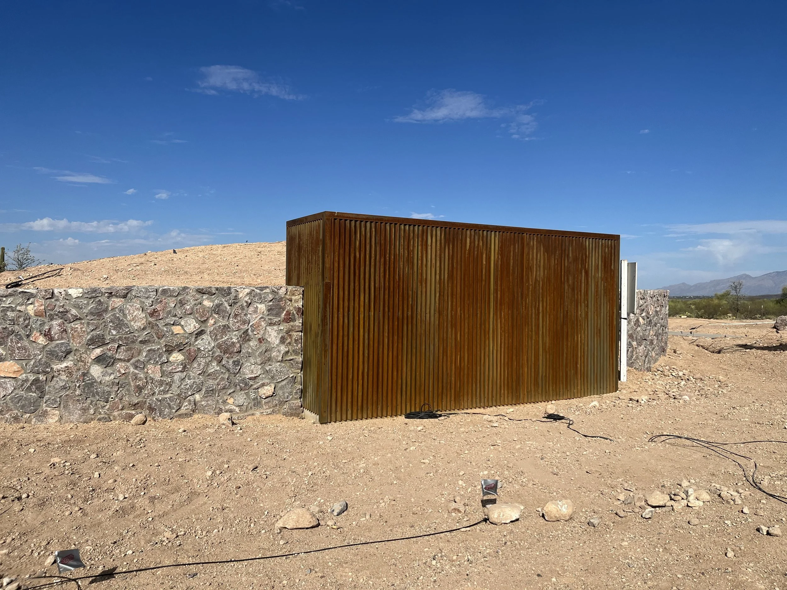 A large wooden block standing on a construction site with a stone wall in the foreground, dirt ground, and a blue sky with some clouds.