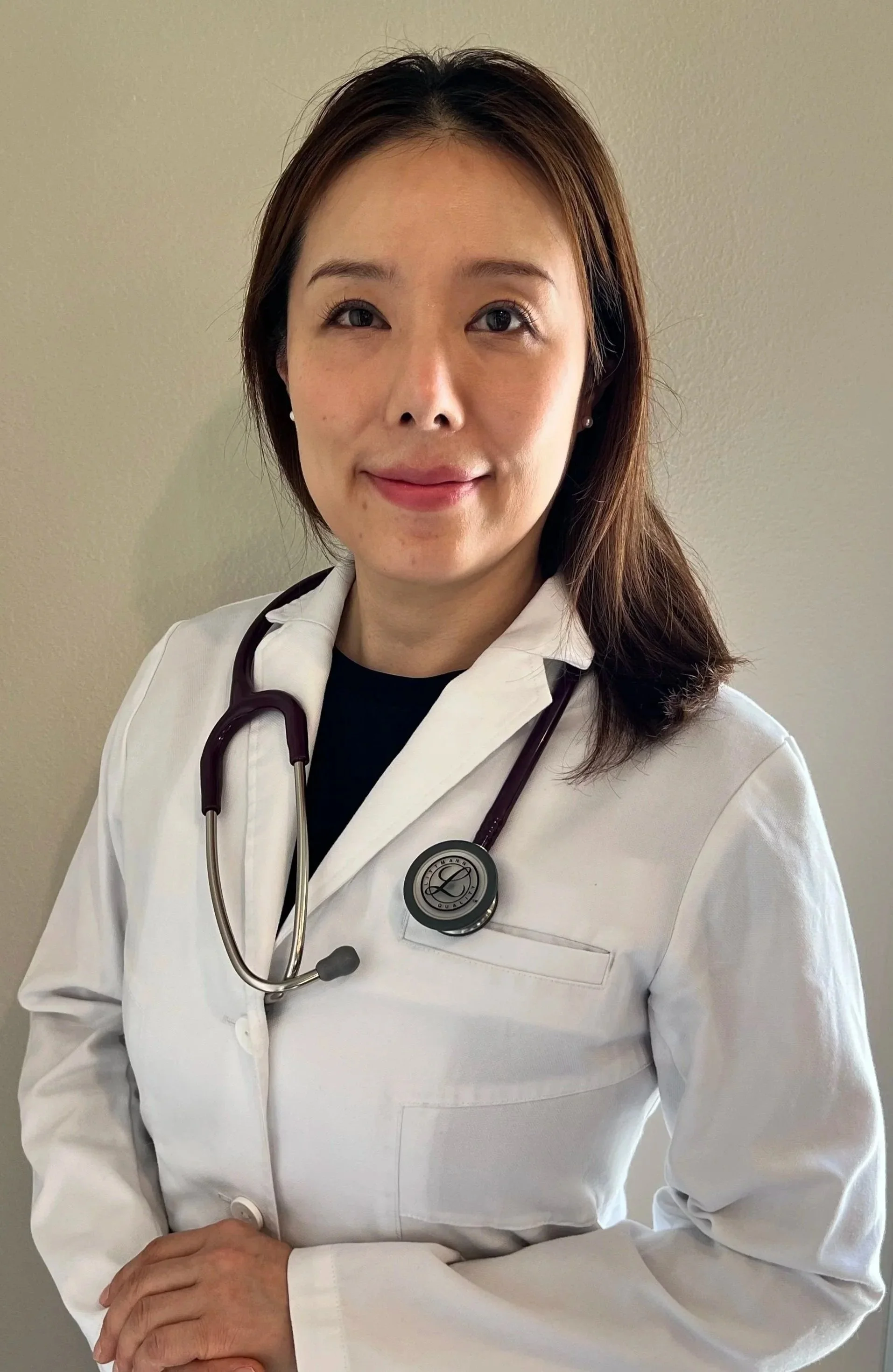 A female doctor wearing a white coat and stethoscope standing against a plain beige wall.