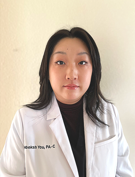 A woman in a white lab coat with black hair standing against a beige wall.