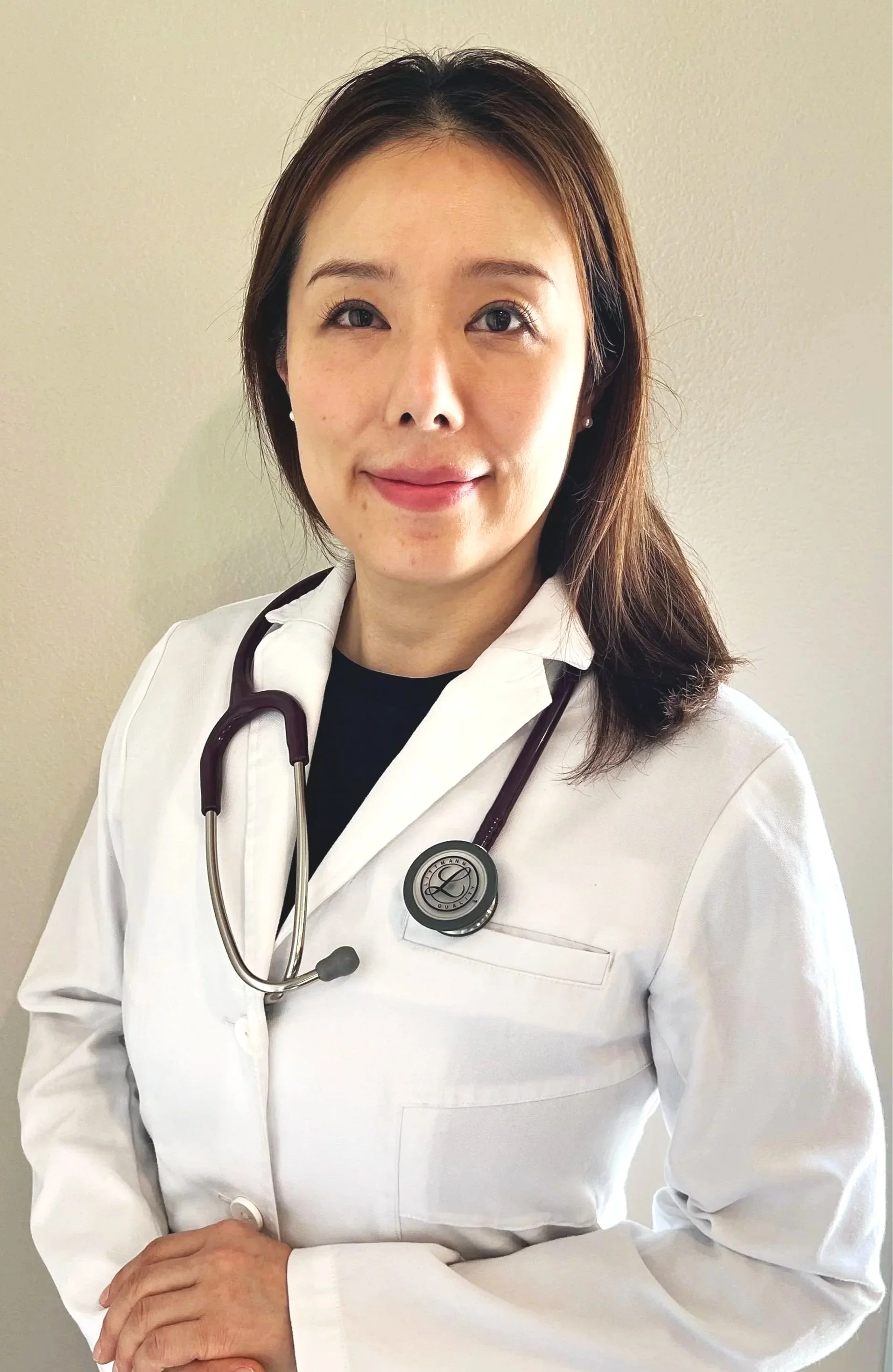 A female healthcare professional wearing a white lab coat with a stethoscope around her neck, standing against a plain wall.