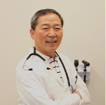 A smiling male doctor with gray hair wearing a white lab coat and a stethoscope around his neck, standing with arms crossed in a clinical setting.