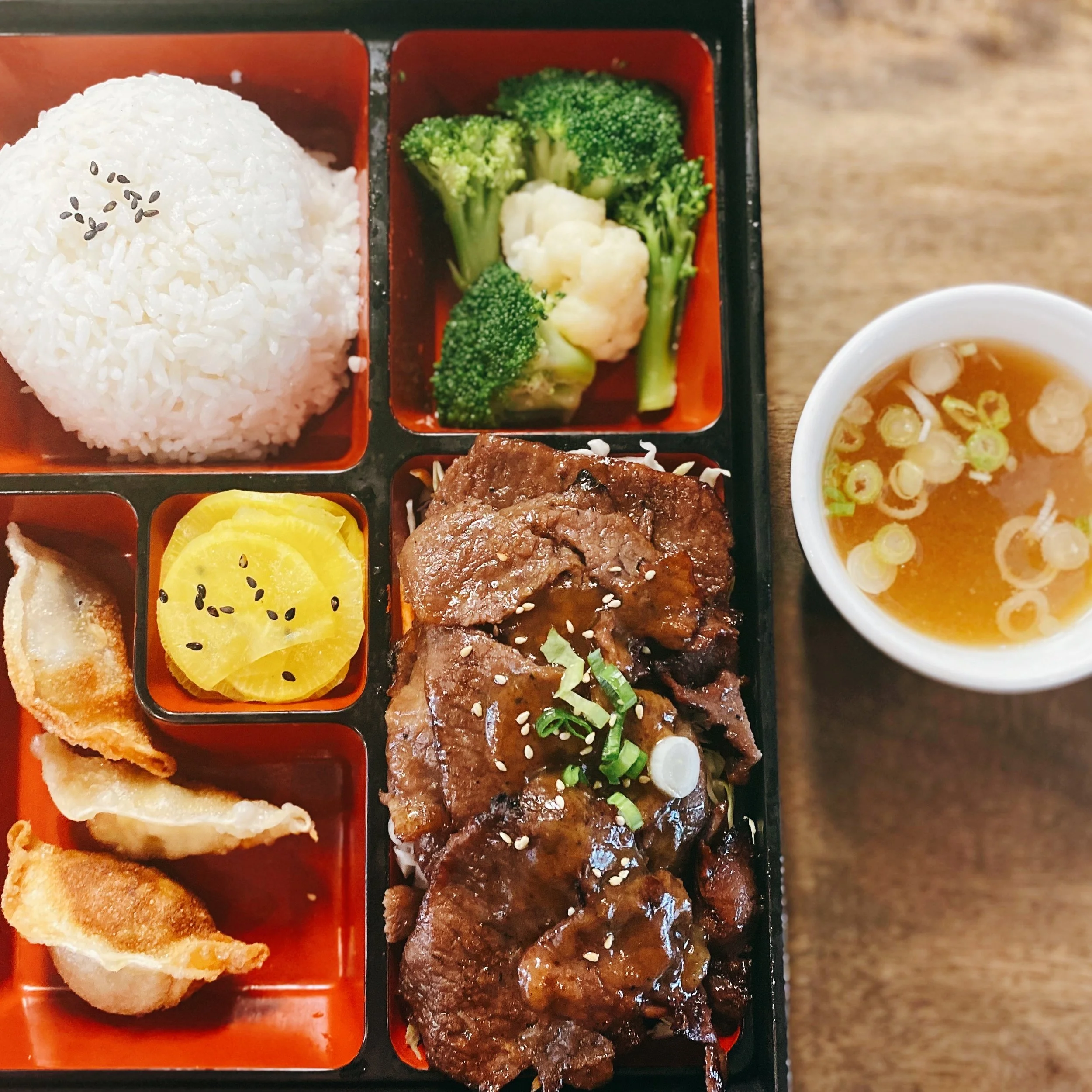 Bento box containing white rice with black sesame seeds, steamed broccoli, cauliflower, sliced yellow pickled vegetables, a piece of fried fish, and beef with green onions, garnished with sesame seeds.