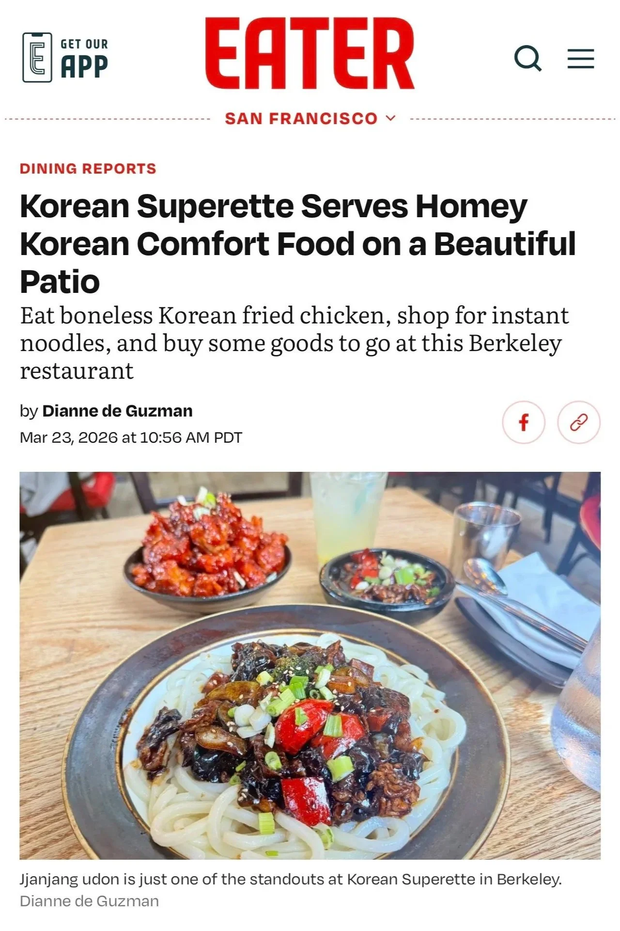 Plate of Korean Jajjang udon noodles topped with vegetables and sauce, with side dishes including spicy red kimchi and chopped vegetables on a eating table.