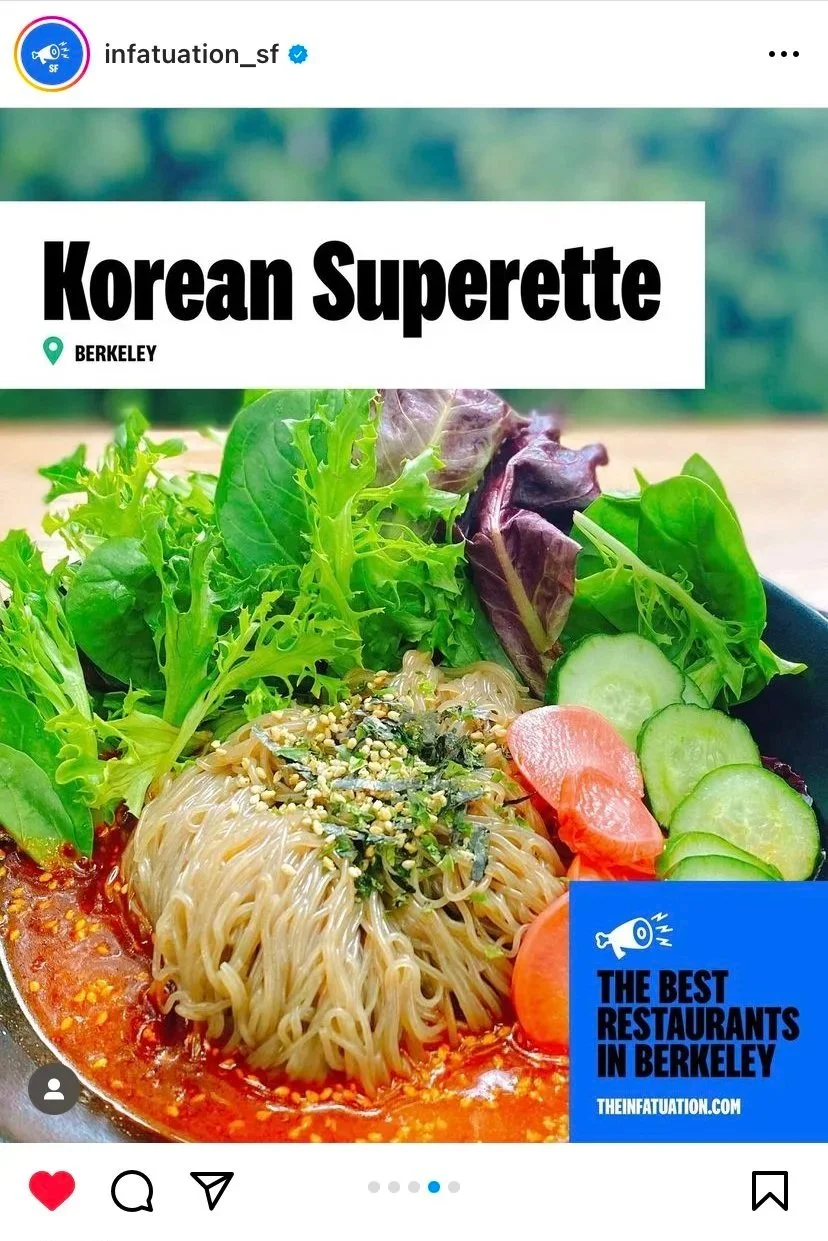 A bowl of Korean cold noodles topped with sesame seeds, served with fresh lettuce, cucumber slices, and carrot slices, with a spicy sauce at the bottom, on a light wooden surface.