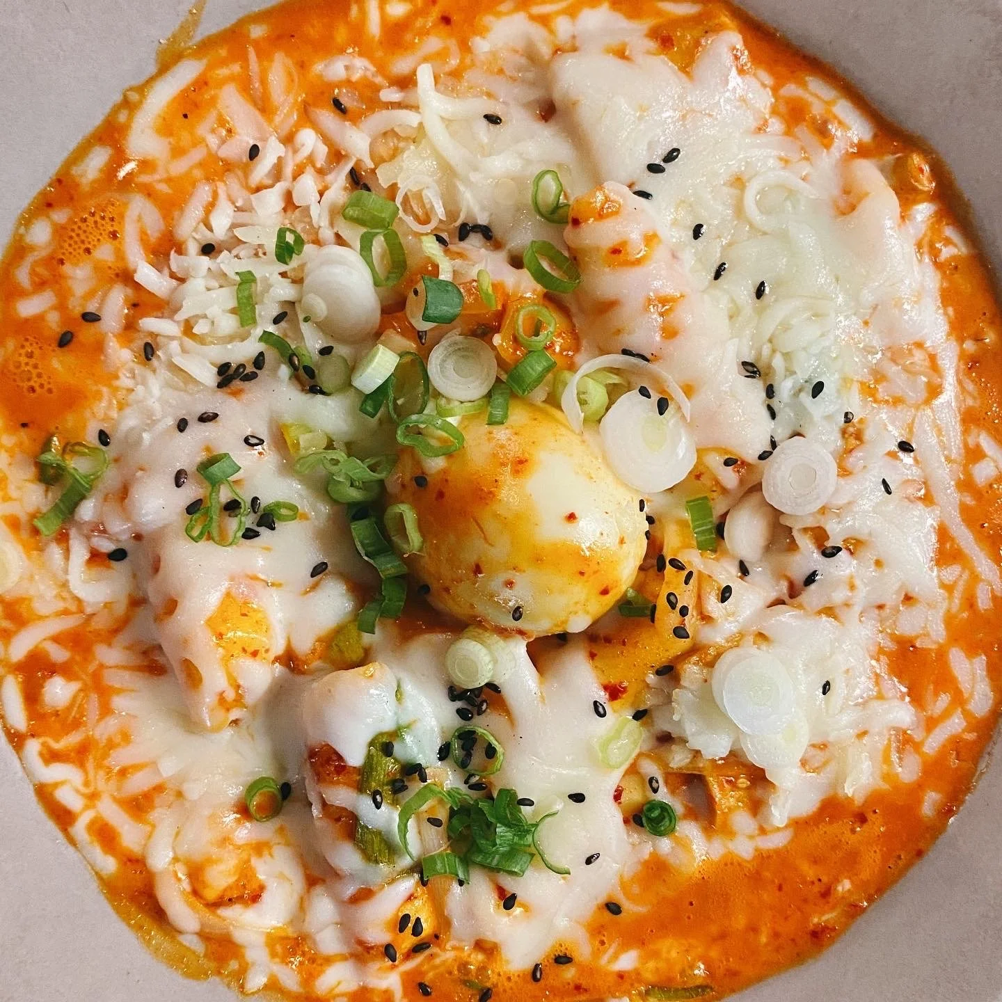 Close-up of a dish featuring melted cheese, chopped green onions, black sesame seeds, and a soft-boiled egg on a spicy orange sauce.