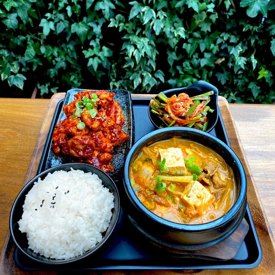 A Korean meal on a black tray with white rice, hot soup with tofu and vegetables, spicy chicken, and kimchi with green onions, set on a wooden table near green leafy plants.
