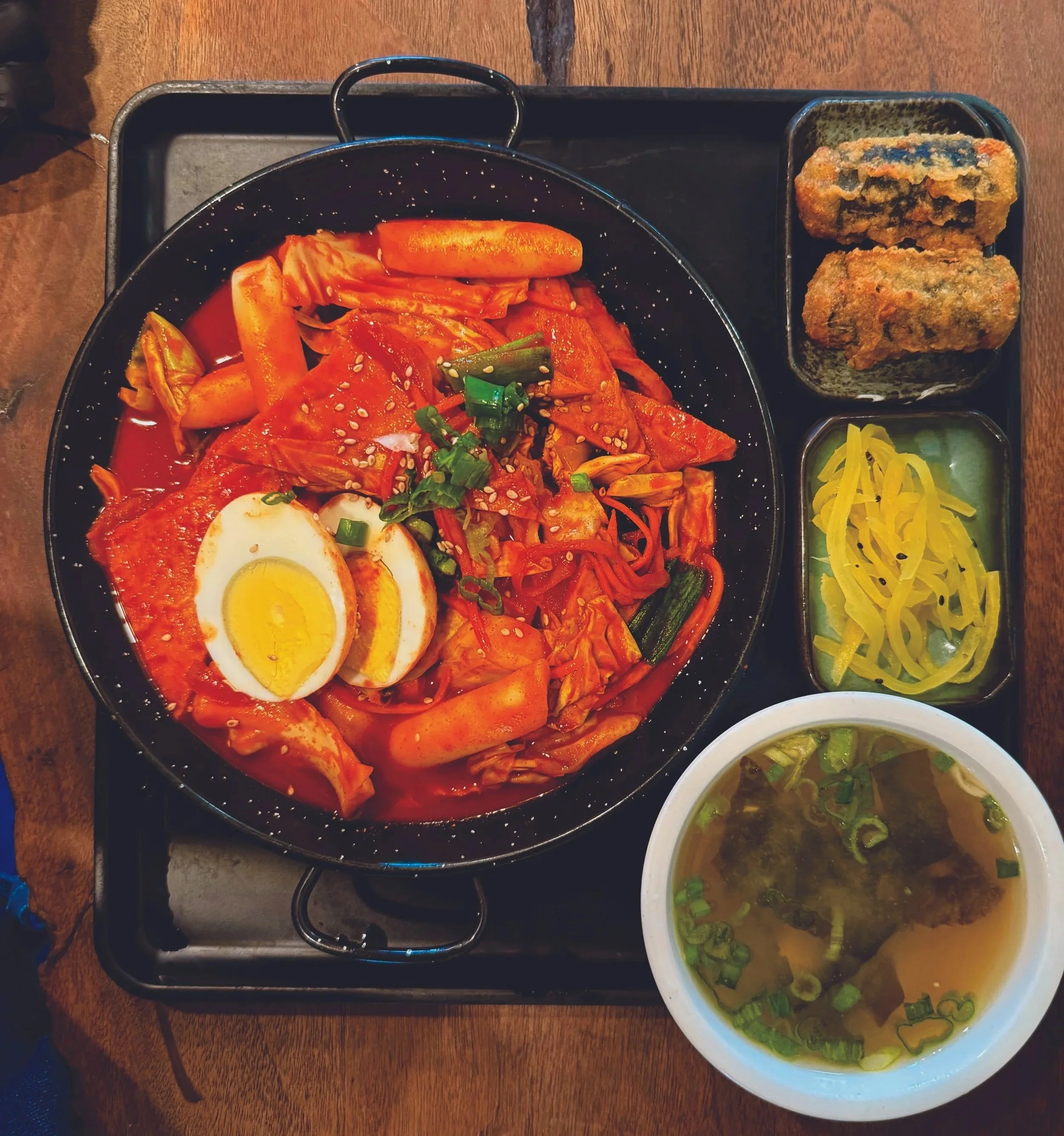 Korean meal with spicy kimchi, boiled eggs, and assorted vegetables in a bowl, two fried vegetable rolls, and side dishes of pickled yellow radish and green onion soup on a wooden table.