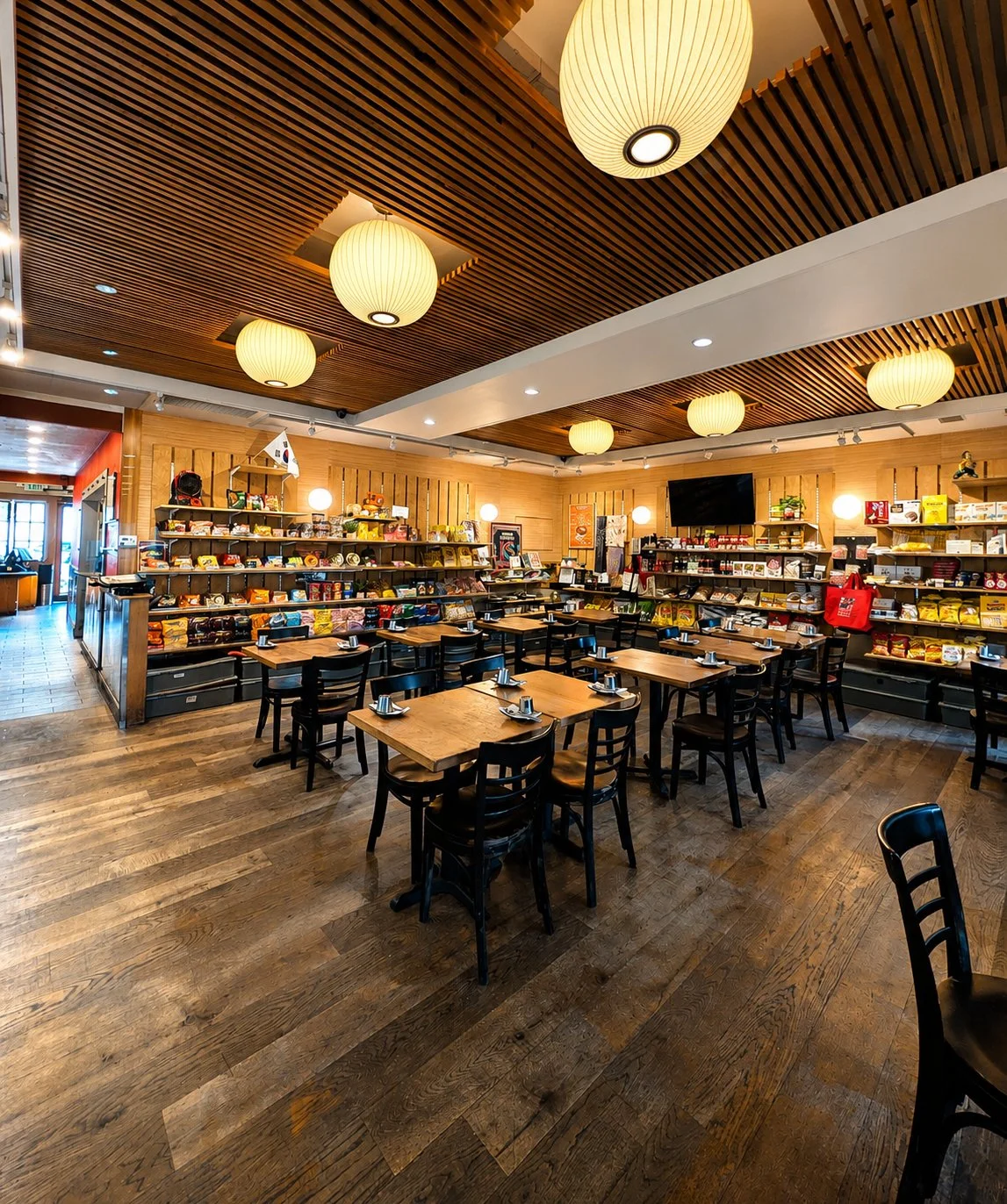 A cozy restaurant with wooden floors and ceiling, featuring round paper lanterns and black chairs around wooden tables. Shelves stocked with snacks and food items line the back wall, with a TV mounted above.