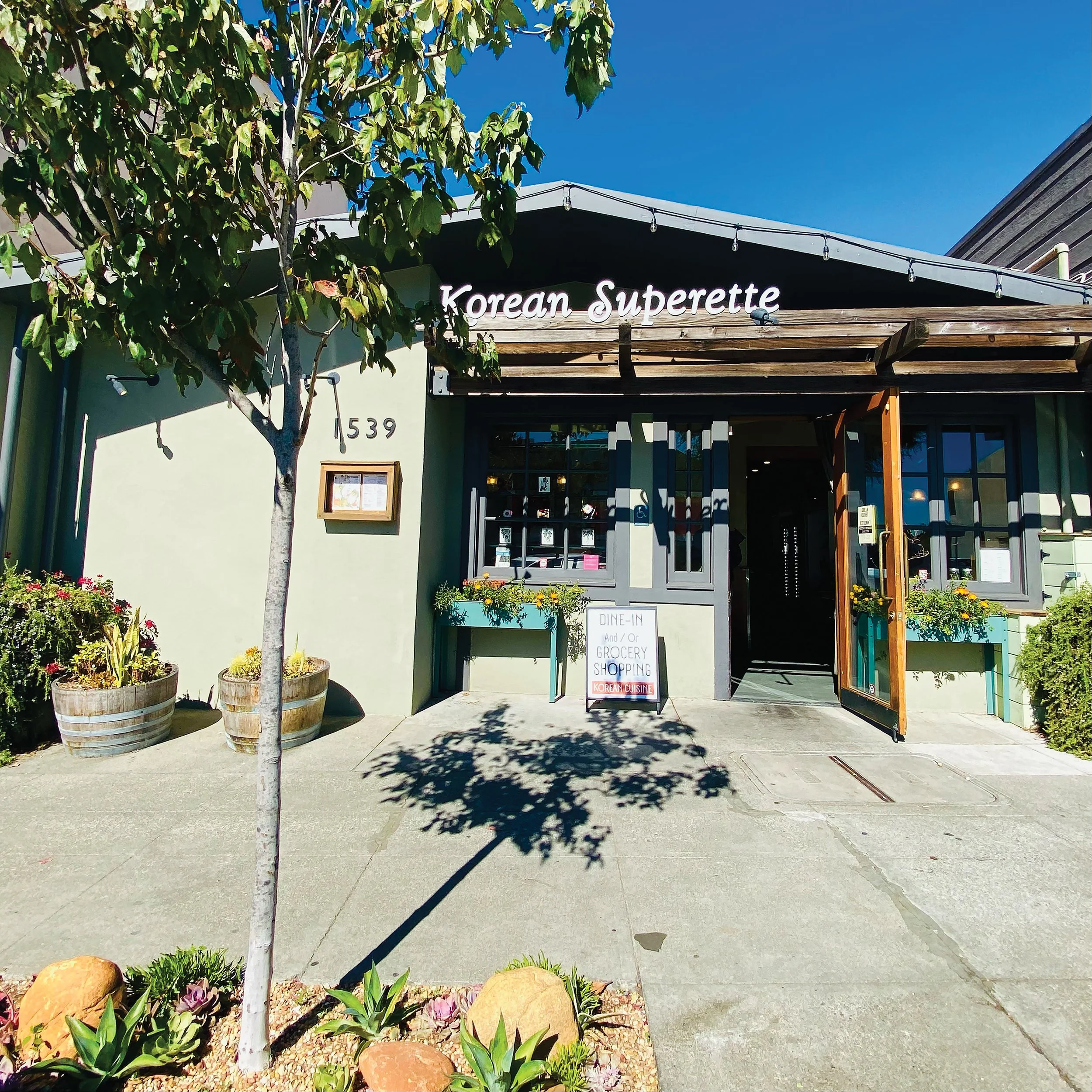 The image shows the front of a restaurant named 'Korean Superette'. There are some plants and flowers in baskets at the entrance, and a signboard indicating dine-in and grocery shopping options with Korean cuisine. A small tree is on the left, casting a shadow on the sidewalk, with a clear blue sky overhead.