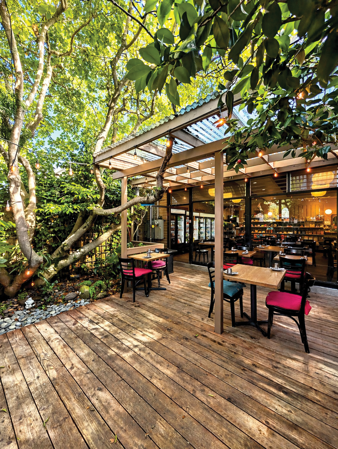 Outdoor patio with wooden flooring, trees, string lights, and tables with chairs in a restaurant or cafe with glass windows showing indoor seating.