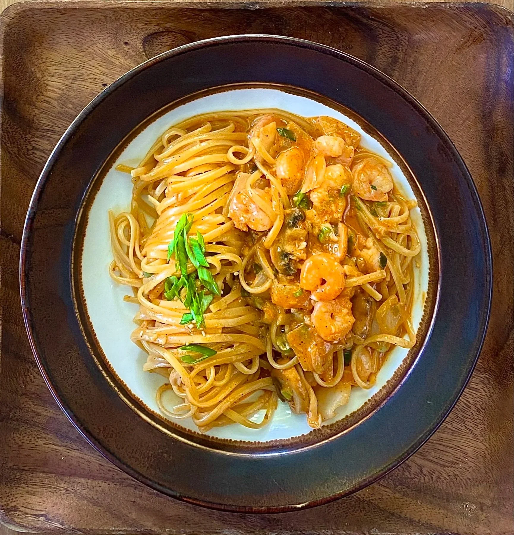 A white plate with a black and brown rim containing a serving of stir-fried noodles with shrimp, vegetables, and herbs, on a wooden table.
