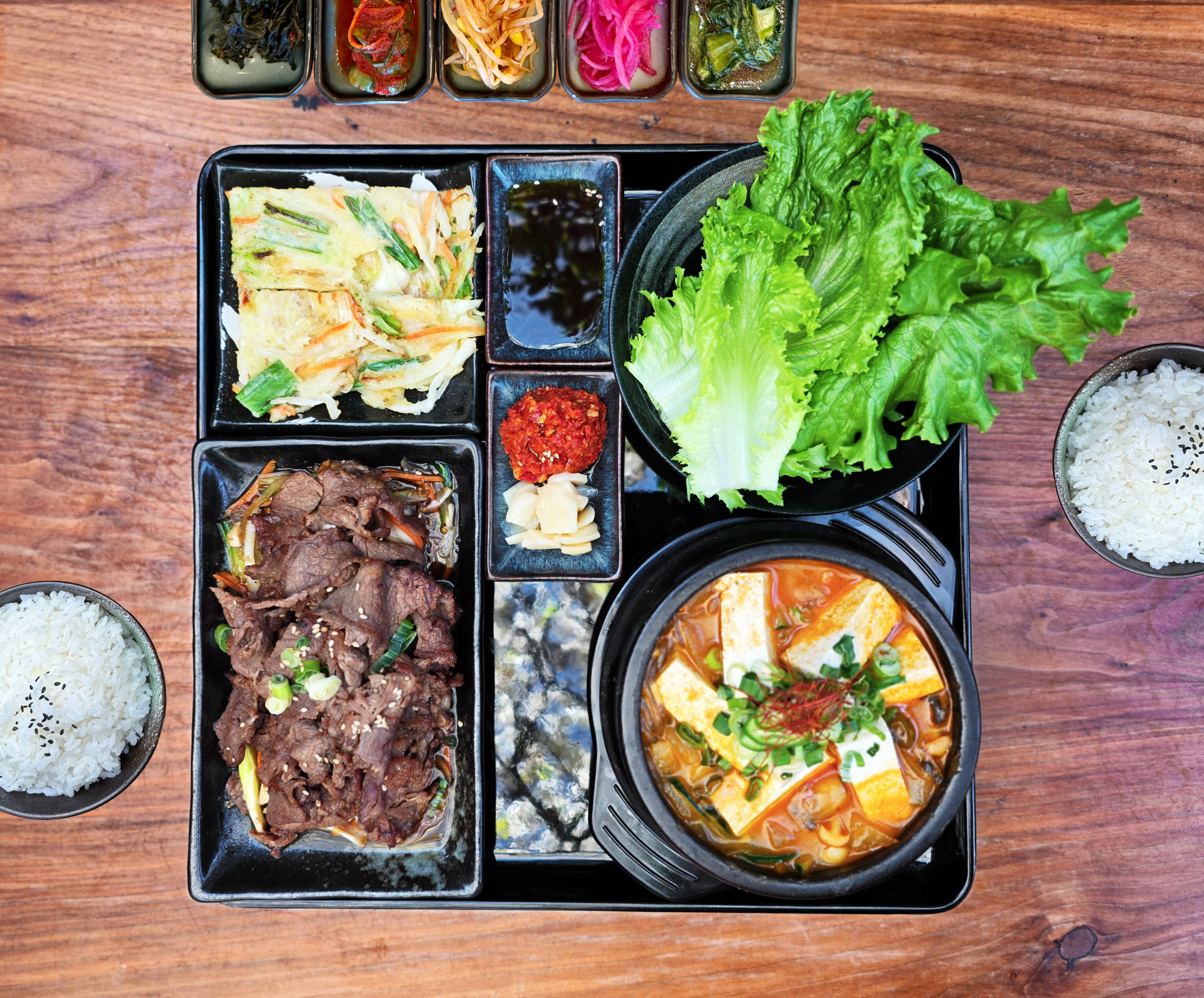 An overhead view of a traditional Korean meal set on a wooden table, featuring multiple small dishes of banchan, a bowl of white rice with sesame seeds, and a pot of spicy stew garnished with green onions and red chili threads.