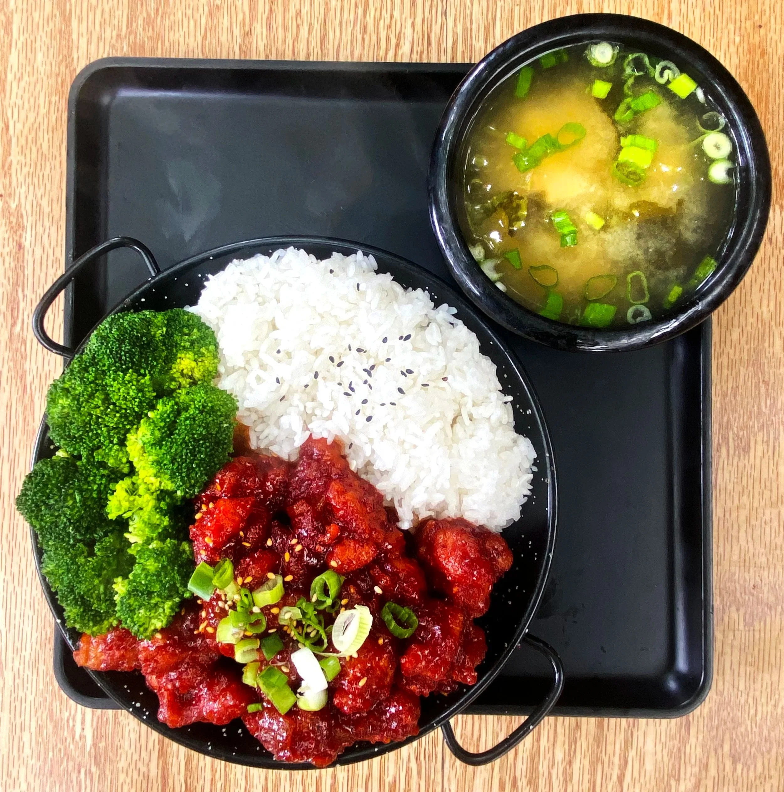 A black tray with compartments containing white rice, broccoli, raw meat in red sauce, and a bowl of miso soup garnished with chopped green onions on a wooden table.