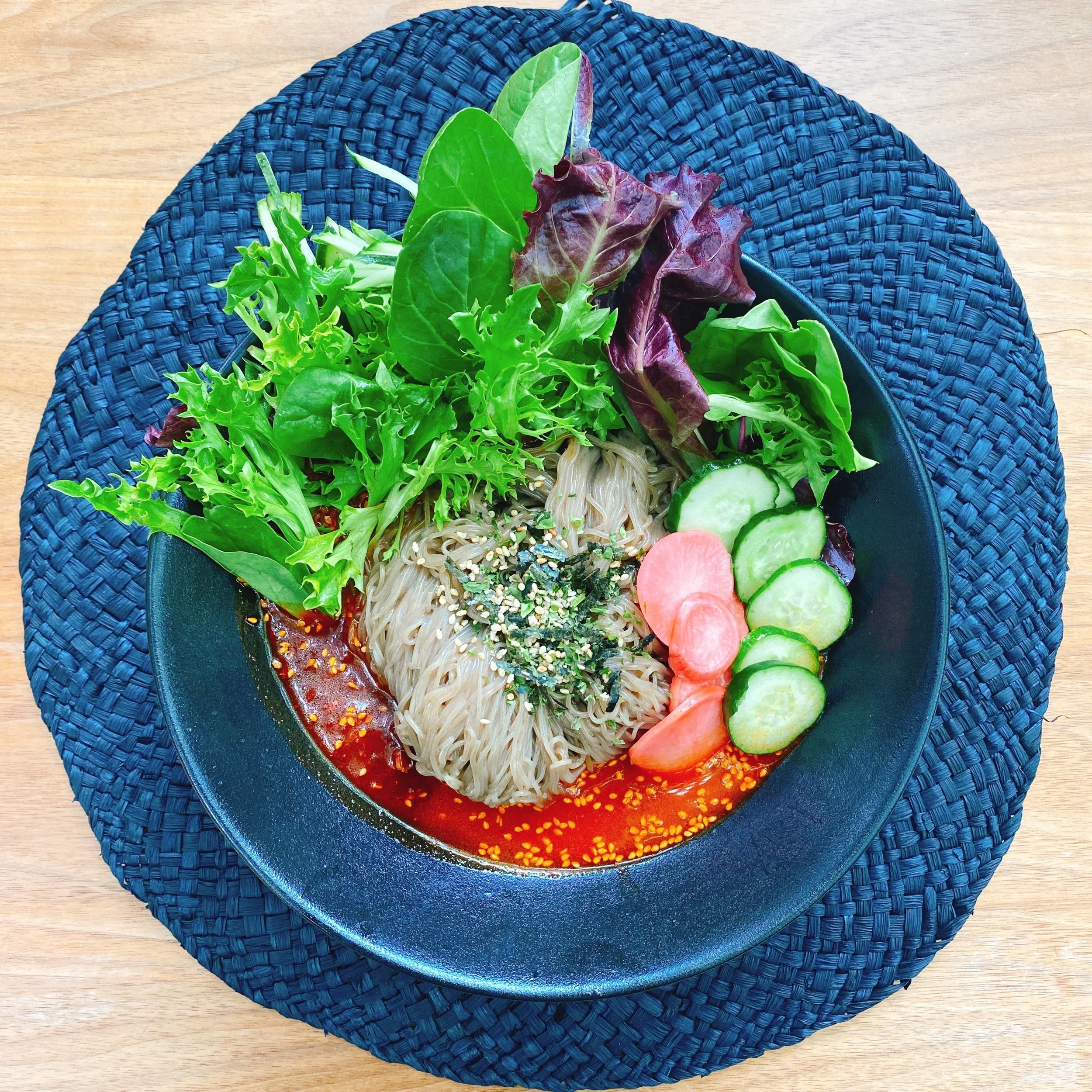 A bowl of assorted fresh greens including lettuce and spinach, with slices of cucumber and radish on a plate, served on a woven blue placemat on a wooden table.