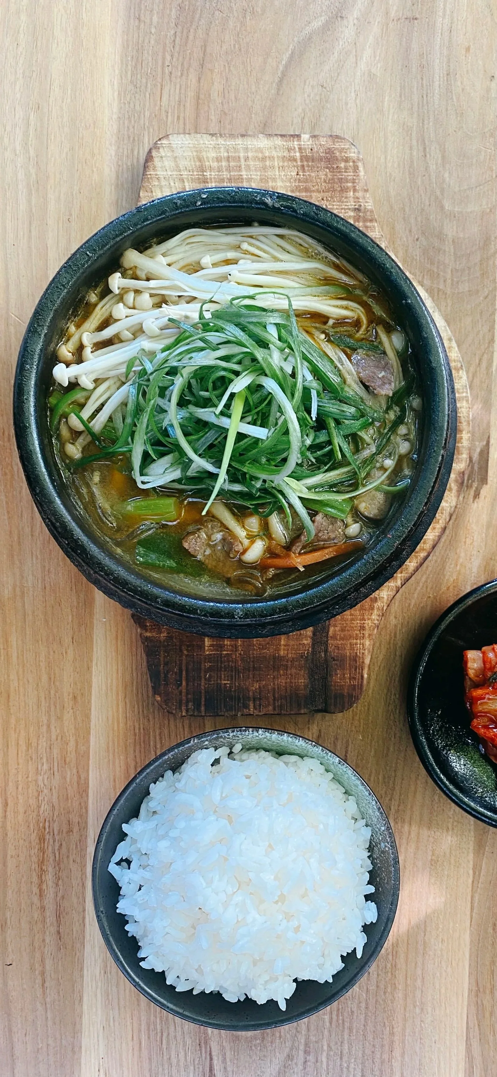 A hot bowl of Korean-style beef stew with vegetables and noodles, served over a wooden board, accompanied by a bowl of steamed white rice and a small serving of side dish on a wooden table.