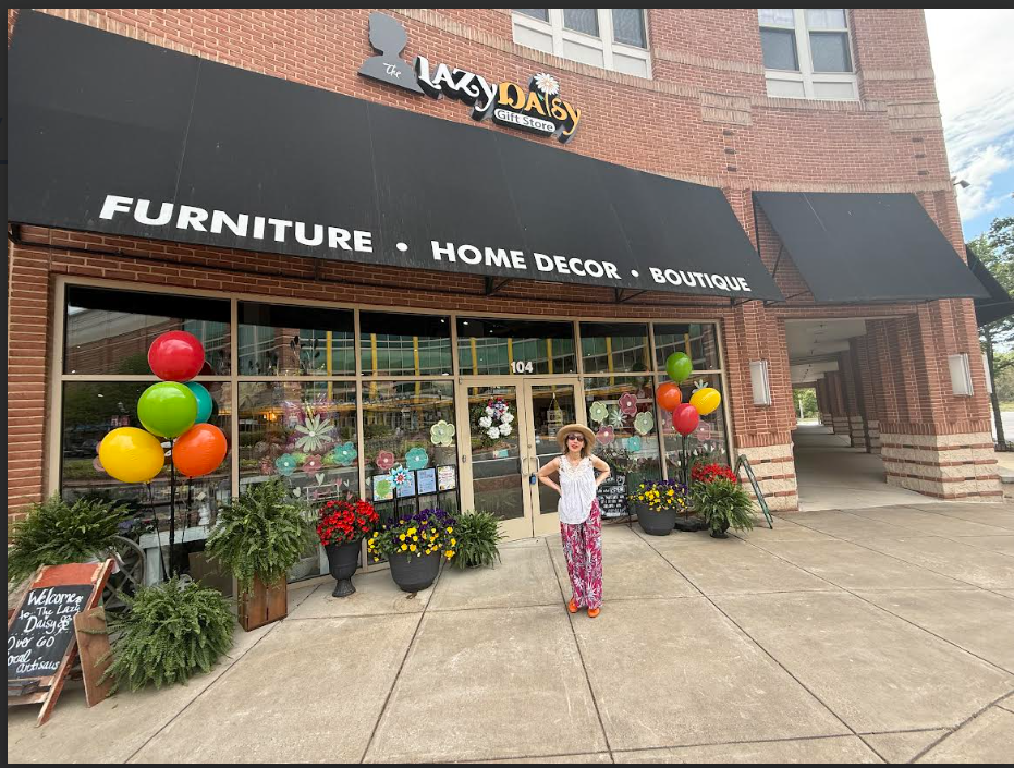 A woman standing outside The Lazy Daisy Gift Store, which sells furniture, home decor, and operates as a boutique. The store entrance is decorated with colorful balloons and potted plants with flowers. The storefront has black awnings and a brick fac