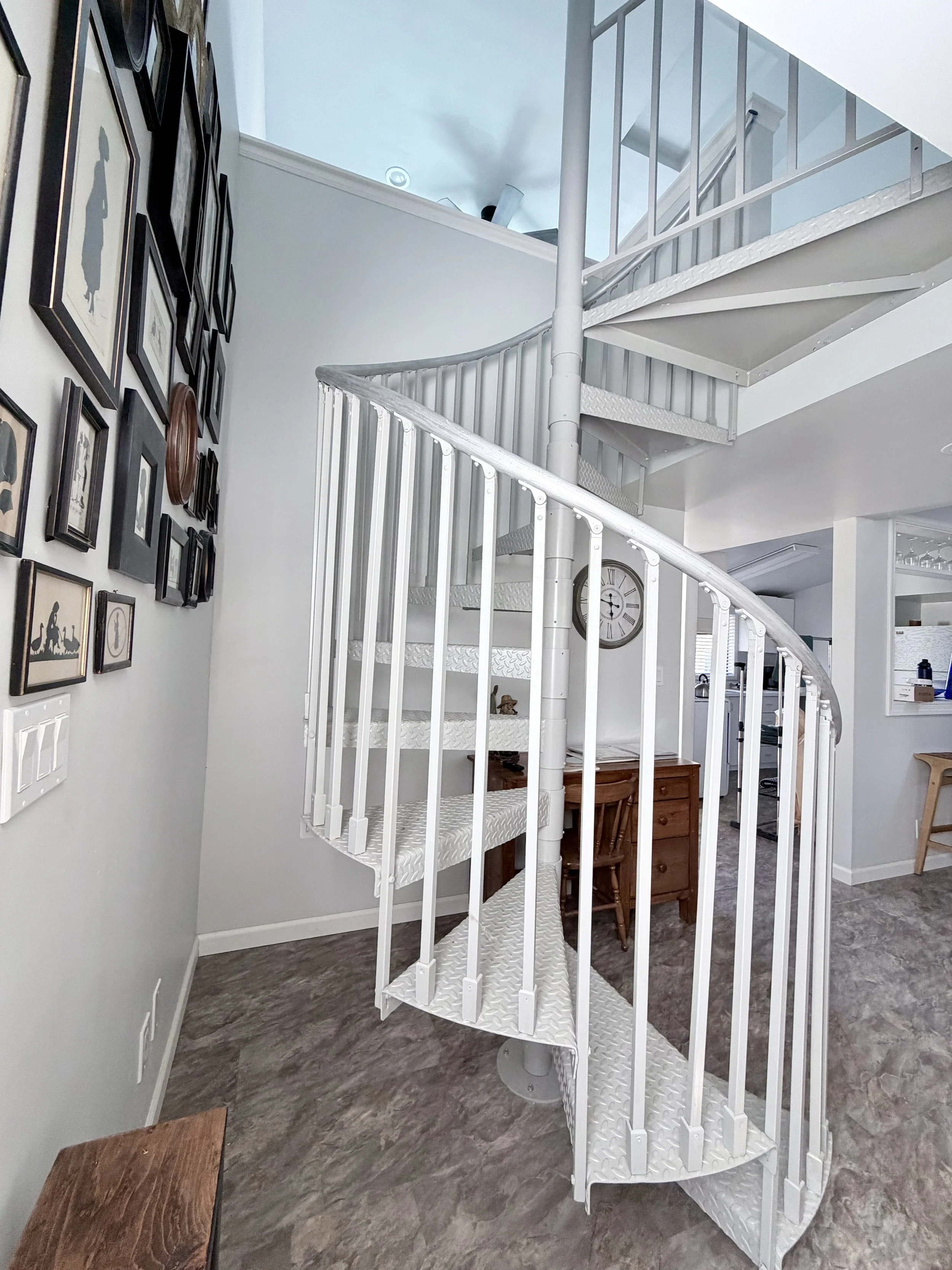 Inside a house showing a white metal spiral staircase with vertical bars, a handrail, and textured steps. The walls are light gray with a picture gallery on one side, and a large wall clock. Part of the kitchen is visible in the background.