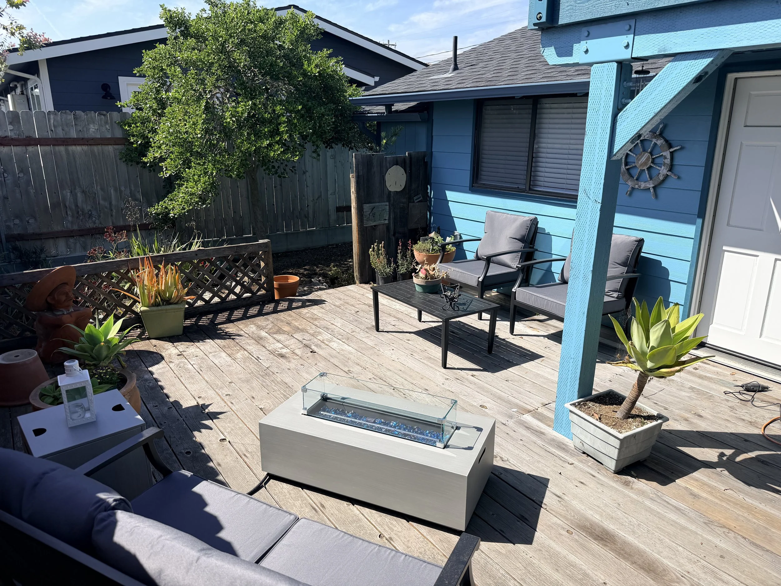 A backyard patio with outdoor furniture, potted plants, and a wooden deck. There is a house with blue exterior walls, a window with closed blinds, and a decorative ship's wheel on the wall. A tree and a wooden fence are in the background, under a clear blue sky.