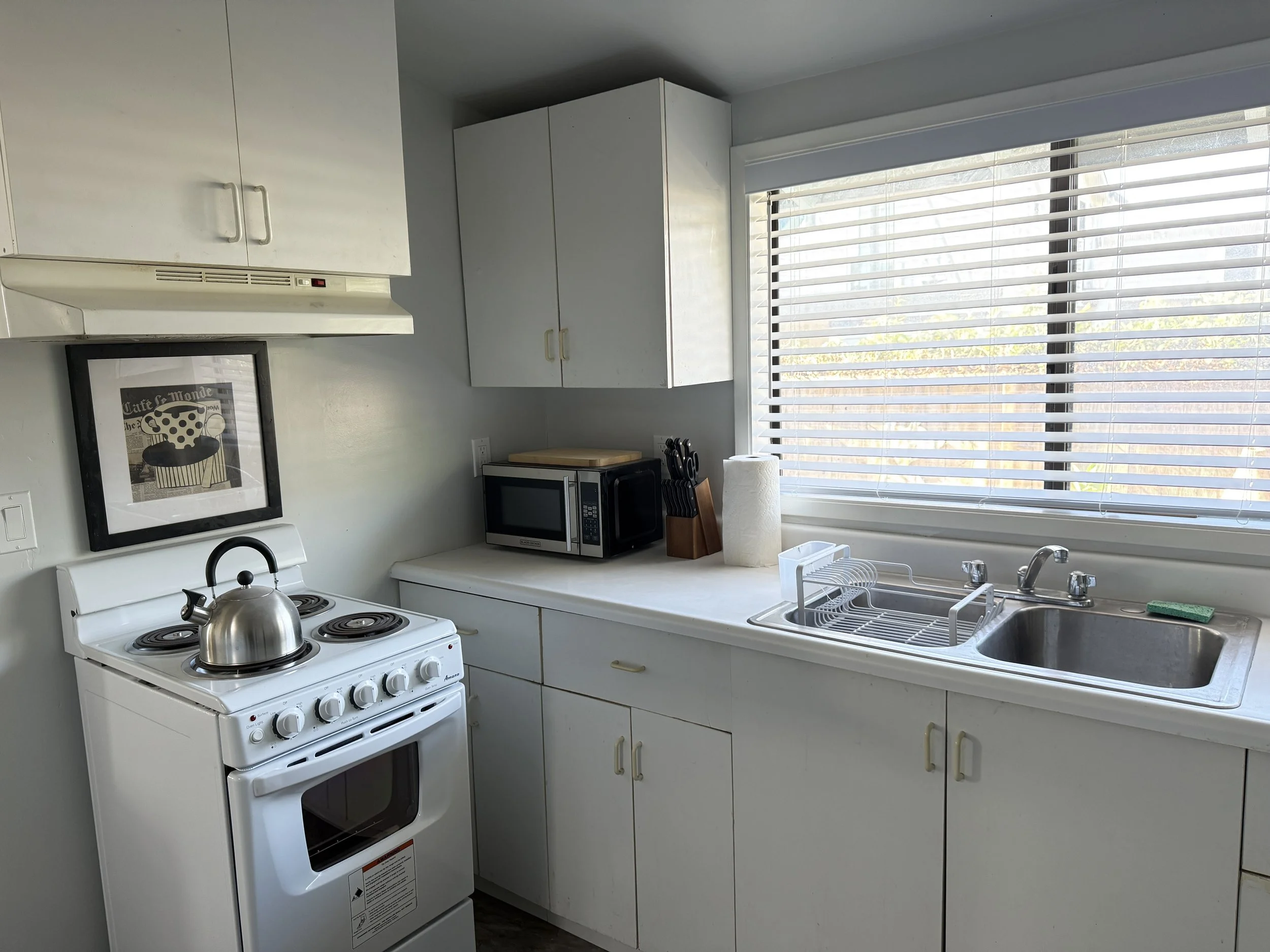 A kitchen with white cabinets, a window with blinds, a stove with a teapot, a microwave, and a paper towel roll on the counter.