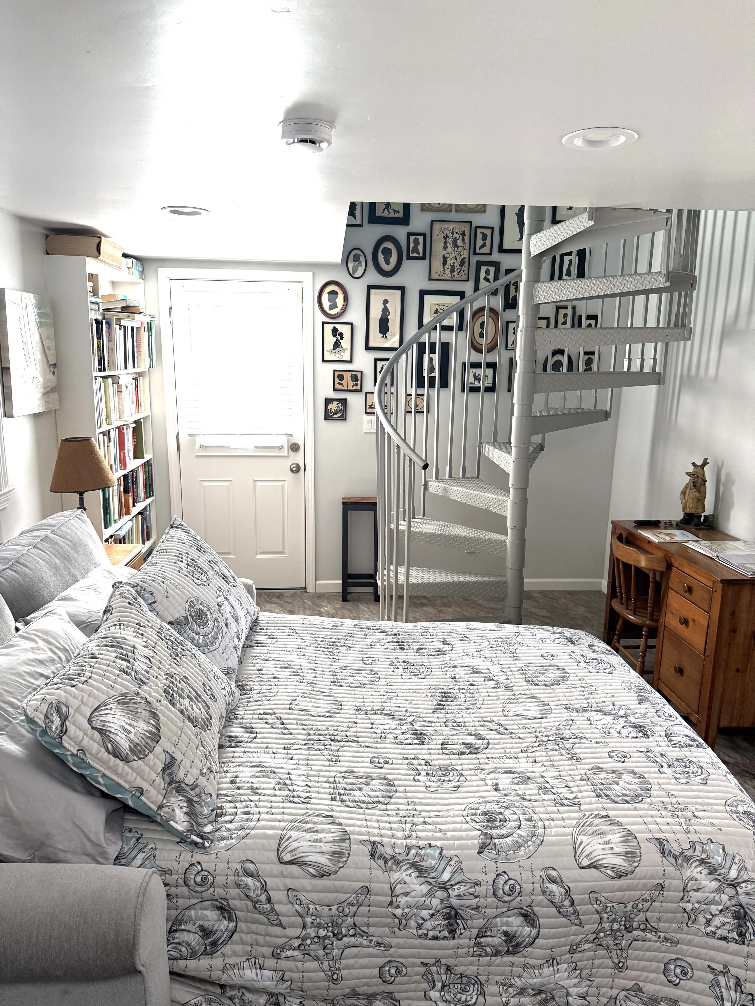 A bedroom with a patterned bedspread and pillows, a bookshelf filled with books, a door with a window, a spiral staircase, and framed wall art.