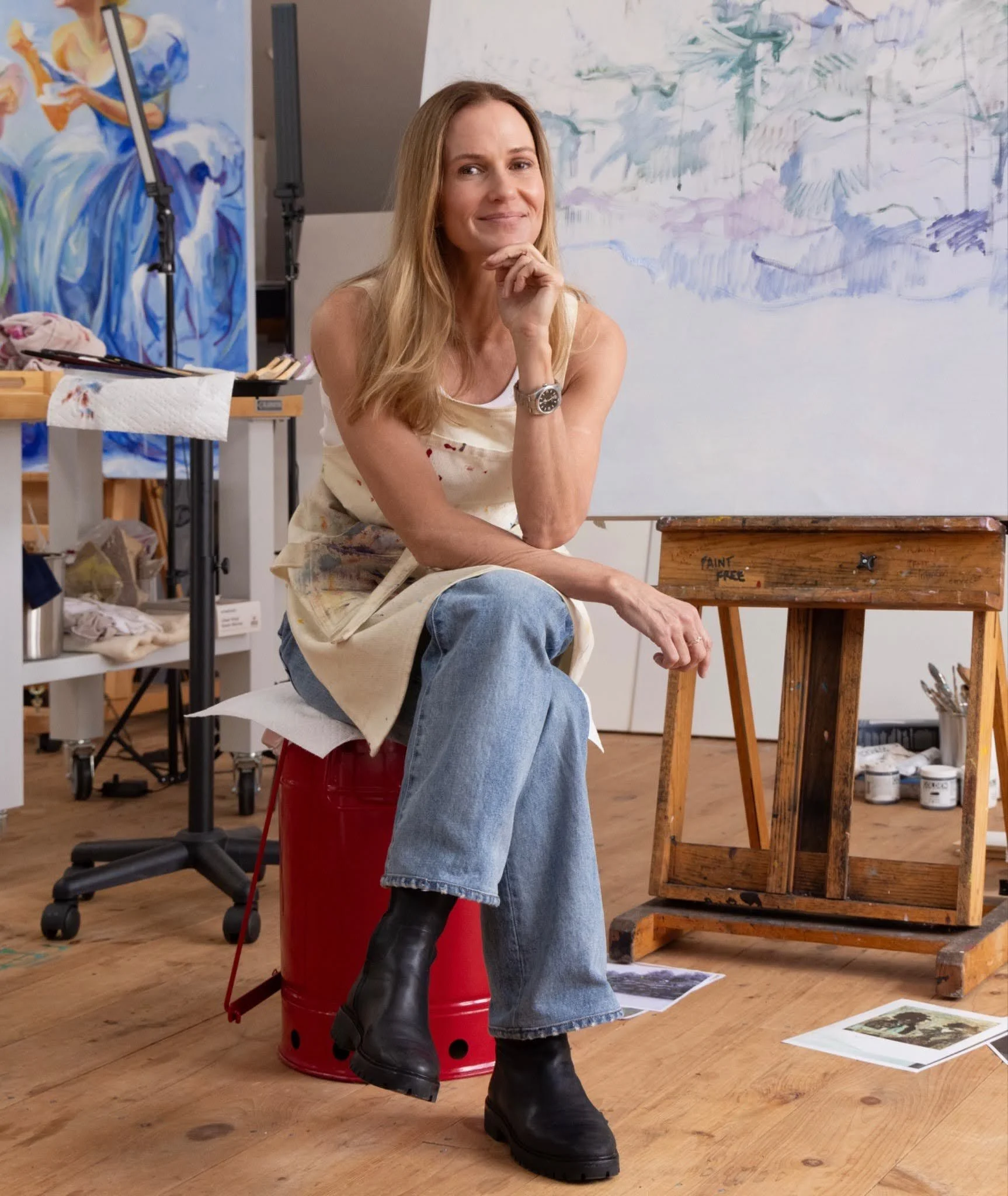 A woman sitting in an artist's studio with paintings and art supplies around her, wearing a sleeveless top, jeans, and large black boots, with a painting in progress behind her.