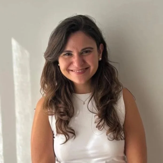 A young woman with long wavy brown hair, wearing a sleeveless white top, smiling at the camera against a neutral background.