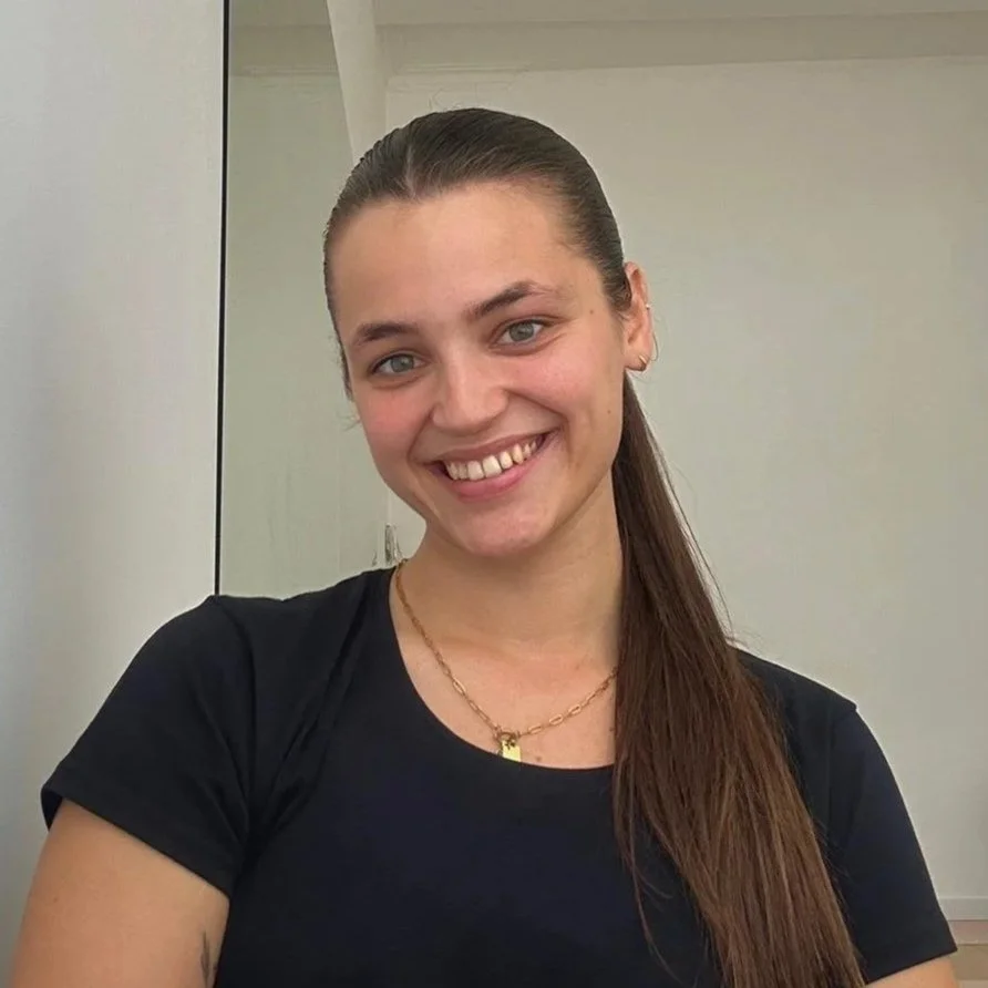 A young woman with long brown hair, smiling, wearing a black t-shirt and a gold necklace, standing indoors against a neutral background.