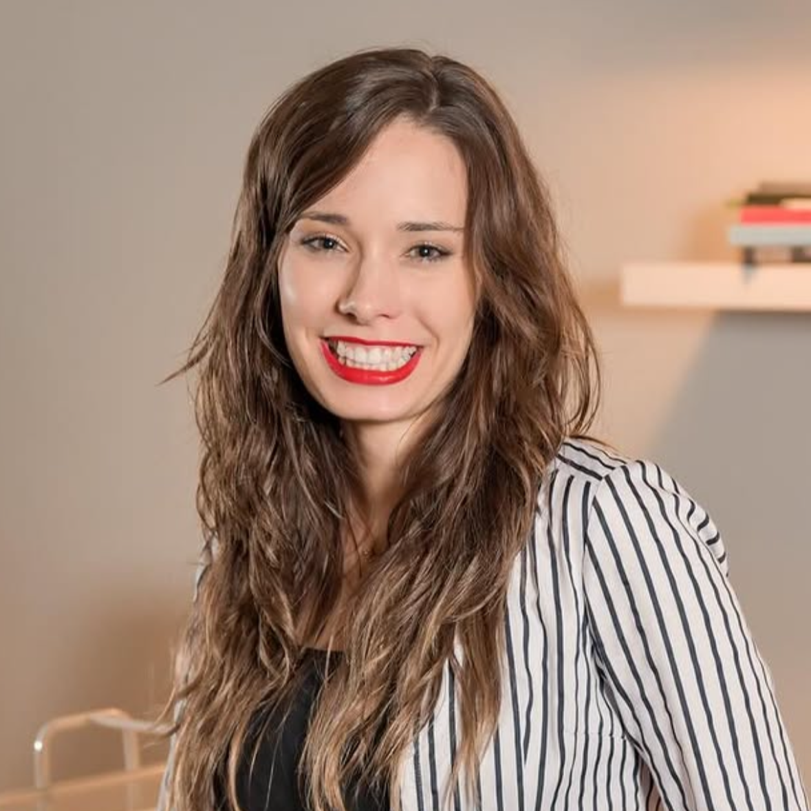 Smiling woman with long wavy brown hair wearing a striped blazer and bright red lipstick in a room with beige walls and a shelf with books in the background.
