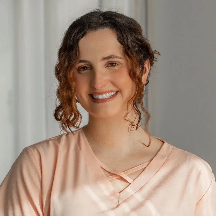 A woman with curly brown hair smiling, wearing a light pink top, standing indoors with a neutral background.