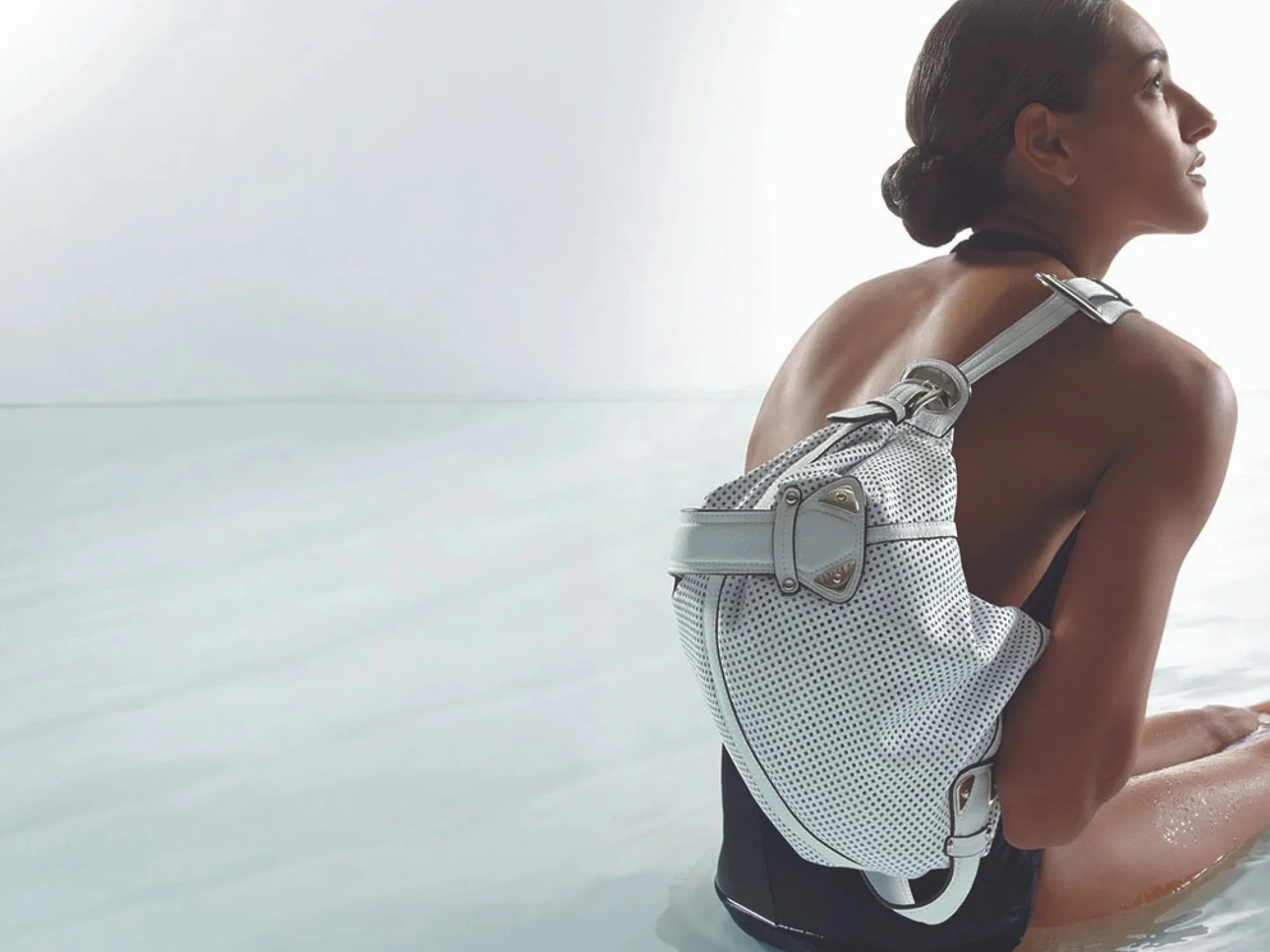 A woman with dark hair in a bun is sitting on the beach, wearing a black swimsuit and a white B.Makowsky shoulder bag with a strap over her shoulder, looking to the right.