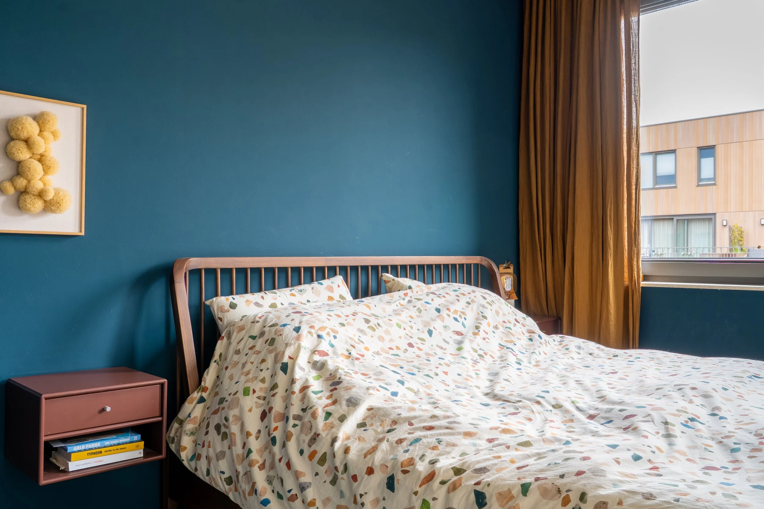 Een slaapkamer met een blauw geschilderde muur, een bed met een matras en een beddengoed met een gekleurd, vlekkerig patroon, een nachtkastje met enkele boeken, een raam met gele gordijnen en een buitengezicht van een modern gebouw.