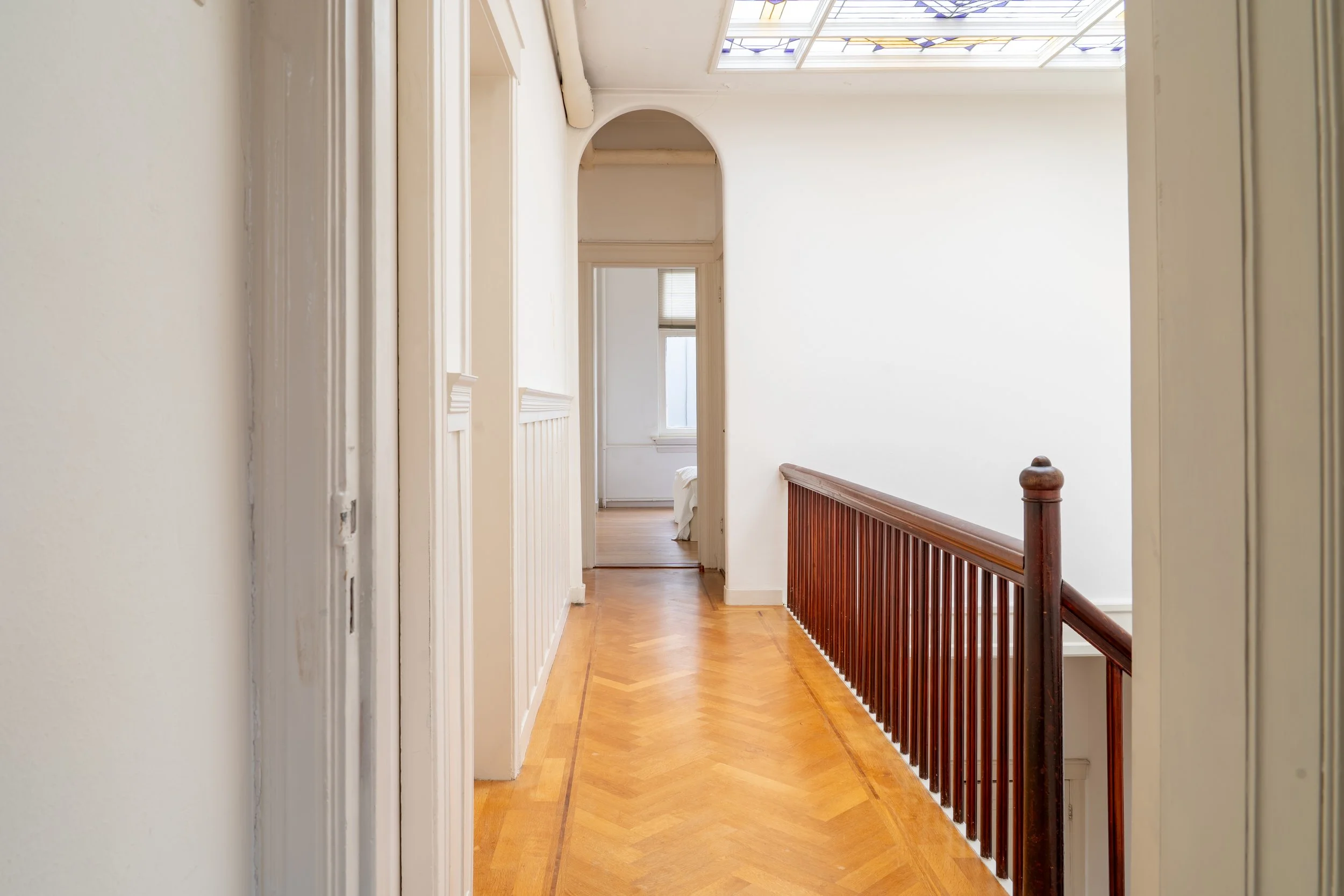 Binnenkant van een huis met houten parketvloer en een houten balustrade, witte muren en ramen met gekleurd glas in het plafond.